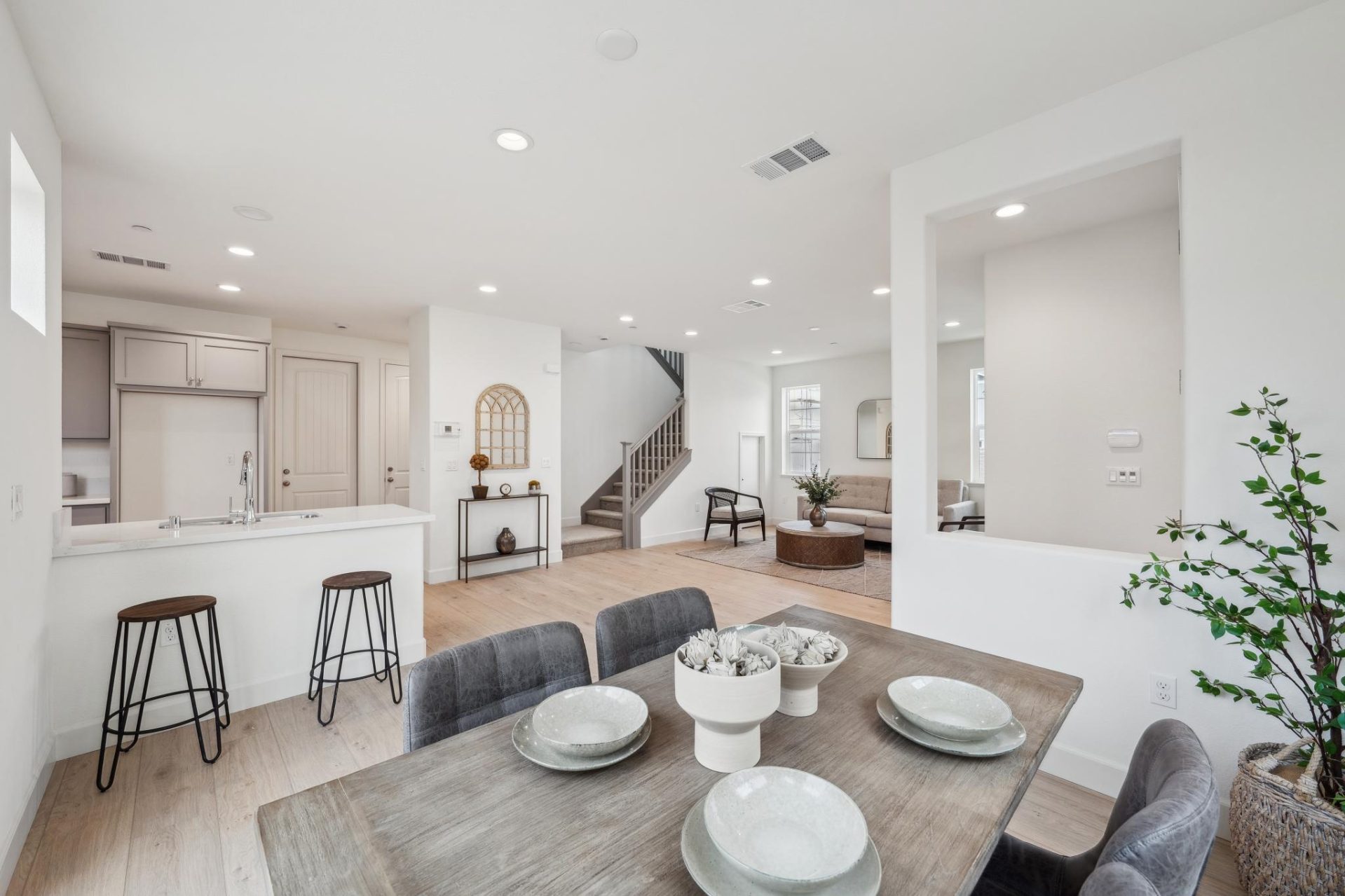 Open-concept main floor with light wood flooring connecting a dining area, kitchen, and living room. A wood dining table with gray upholstered chairs sits in the foreground, the kitchen features gray cabinets and a peninsula with bar stools, and a carpete