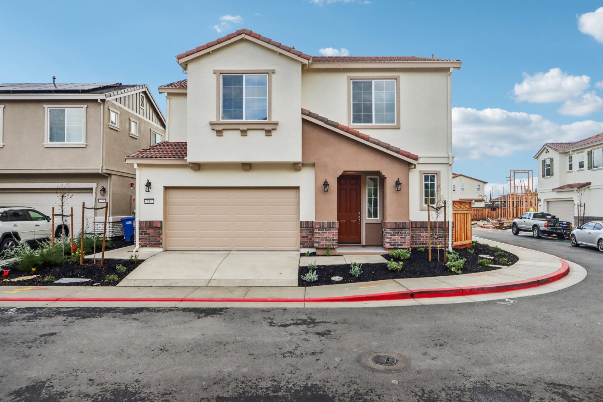 Two-story home with cream and tan stucco exterior, terracotta tile roof, and red brick wainscoting. The home features a two-car garage, a dark wood front door with sidelight, decorative carriage lights, and young landscaping along a corner lot with a red-