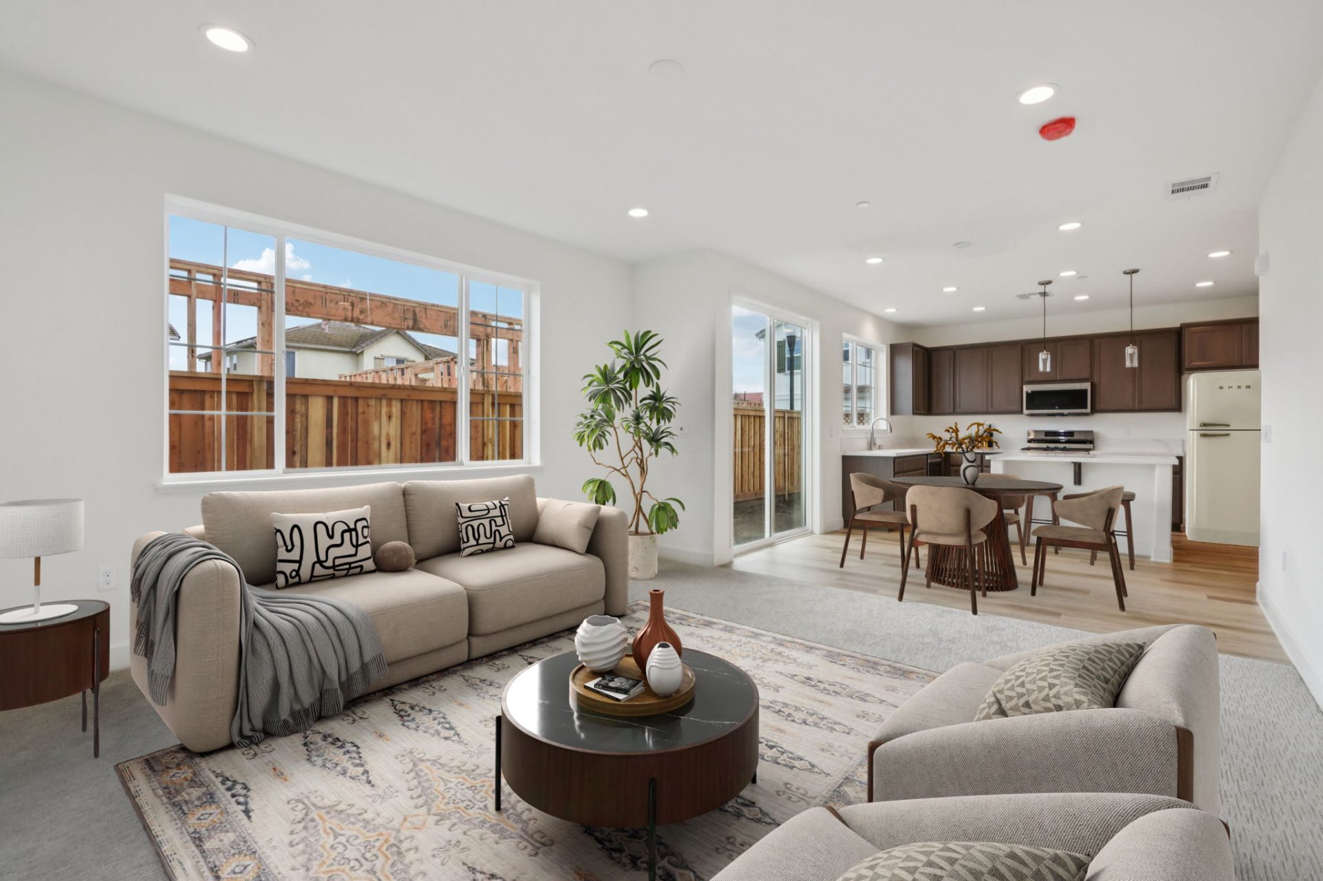 Open-concept living room and kitchen with light walls and wood-look flooring. The living area features neutral sofas with throw pillows, a round wood coffee table, and a potted plant on a patterned rug, while the kitchen beyond has dark wood cabinets, whi