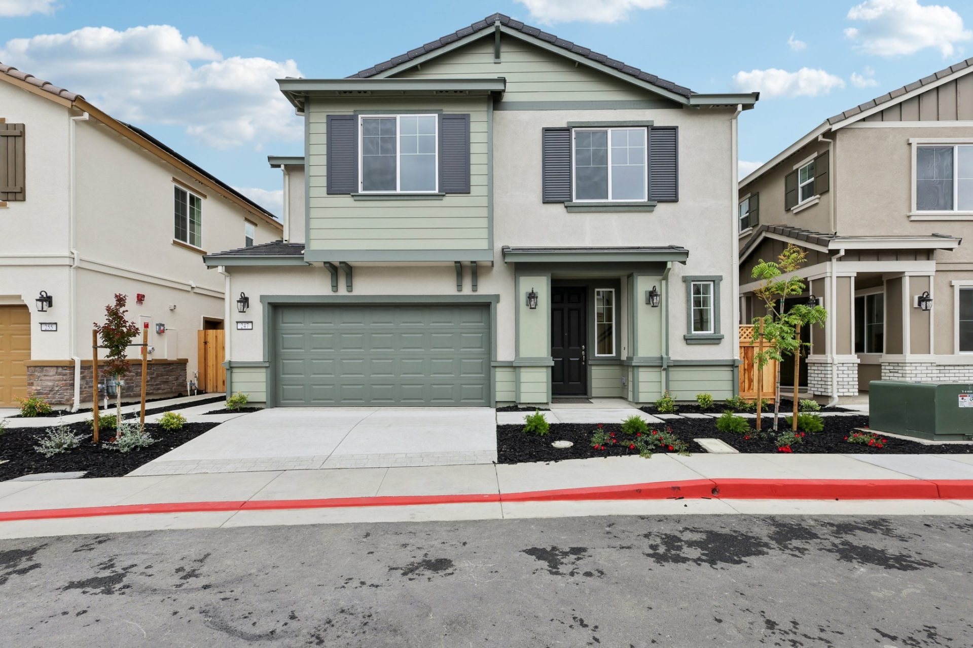 Two-story home with sage green siding and contrasting darker trim, featuring a gabled roofline and a two-car garage with a matching green door. The home has a covered front entry, mixed siding textures including board-and-batten accents, and young landsca