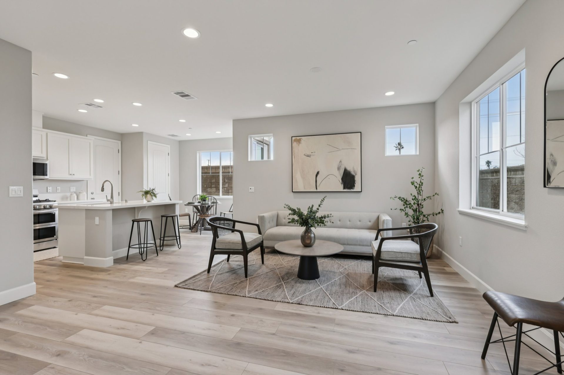 Open-concept living space with light wood-look flooring and gray walls connecting a living room, kitchen, and staircase. Open-concept living space with light wood-look flooring and gray walls connecting a living room, kitchen, and staircase. The foregroun