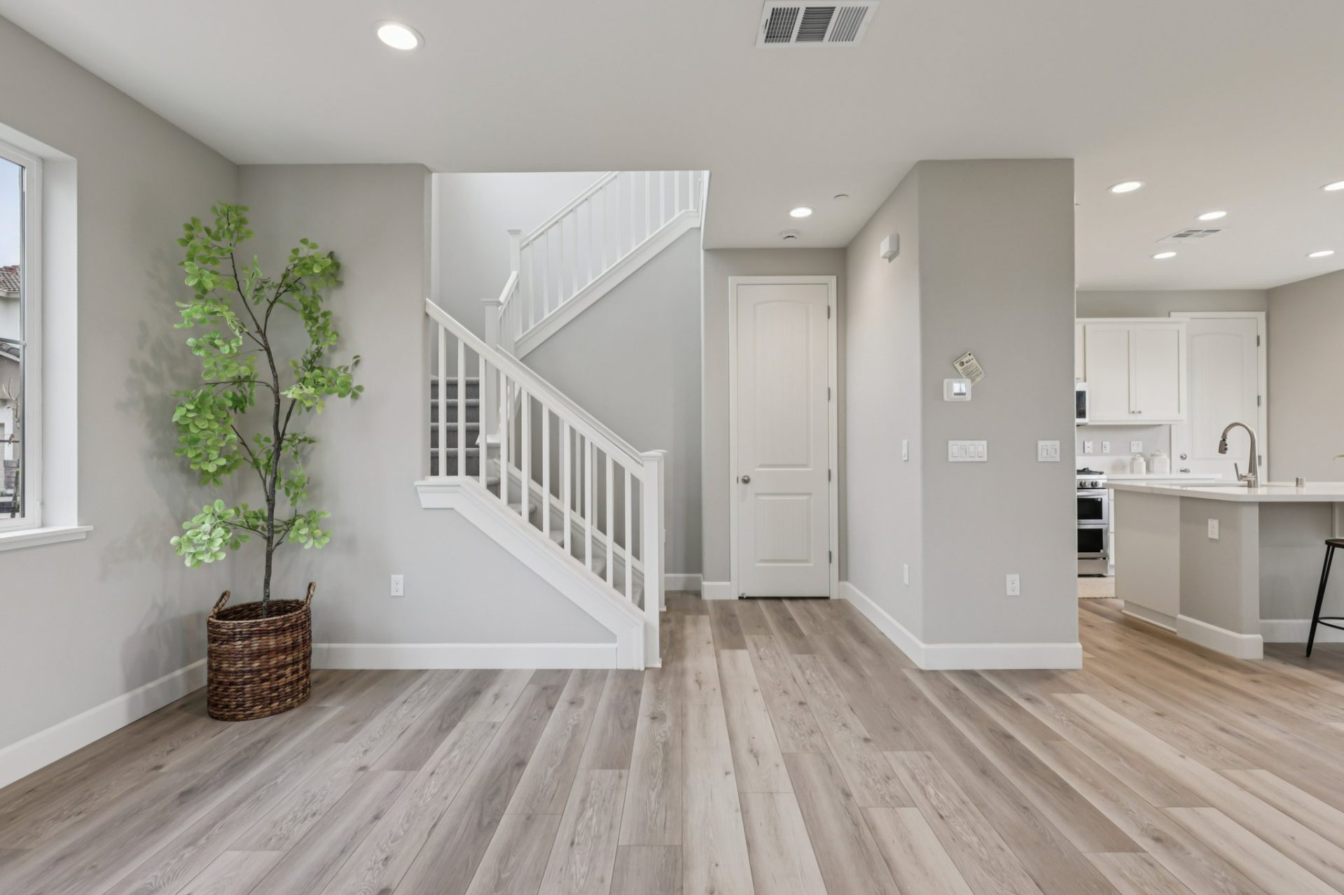 Entryway and staircase area with light gray walls and wood-look flooring. A carpeted staircase with white railings rises along the wall, with a potted tree in a woven basket nearby and a white door under the stairs. The kitchen is visible to the right wit