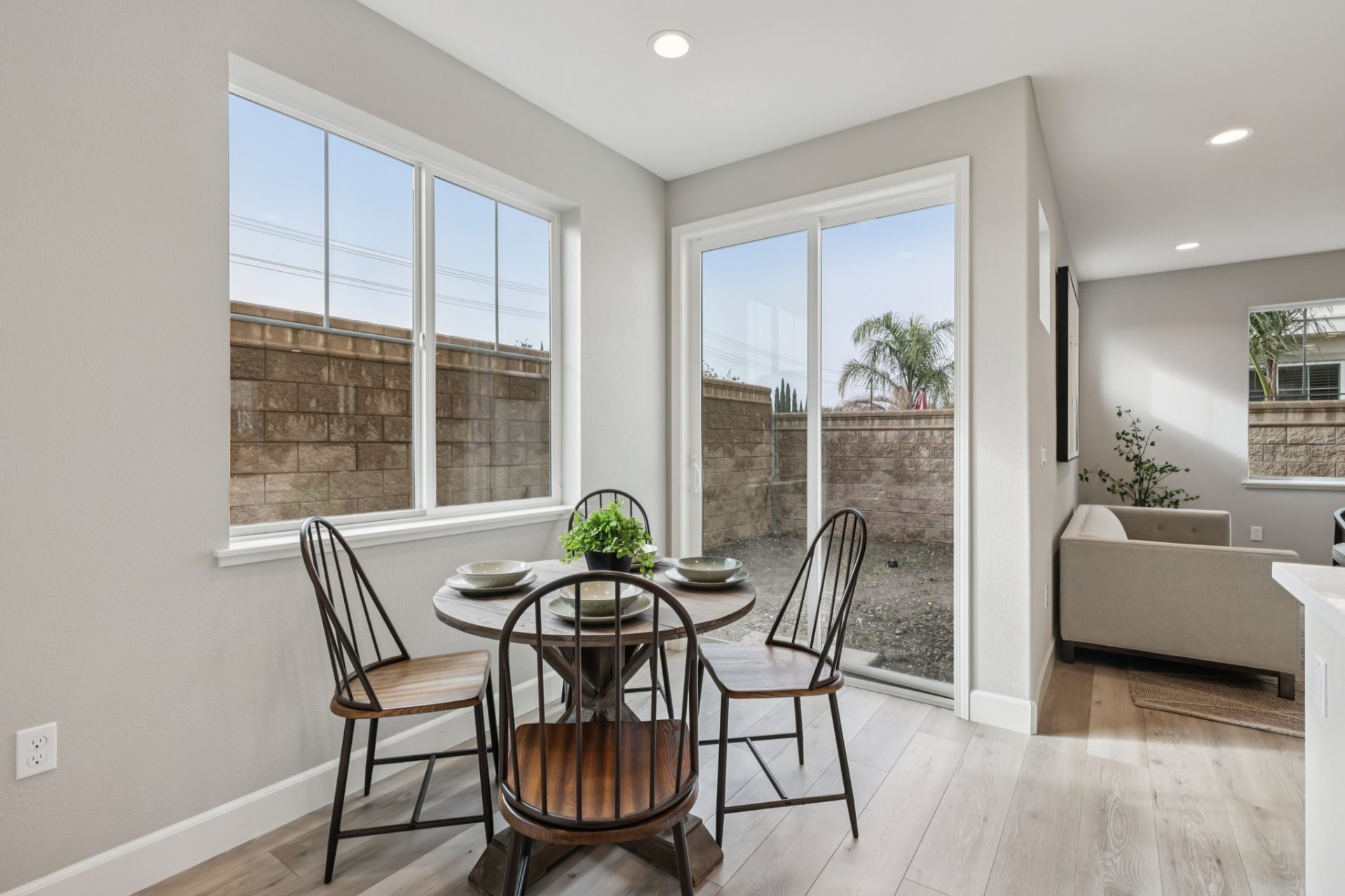 Breakfast nook with a round wood dining table and Windsor-style chairs, set beside large windows and a sliding glass door. The space has light gray walls and wood-look flooring, with natural light coming in from the backyard view of a block wall fence. Th