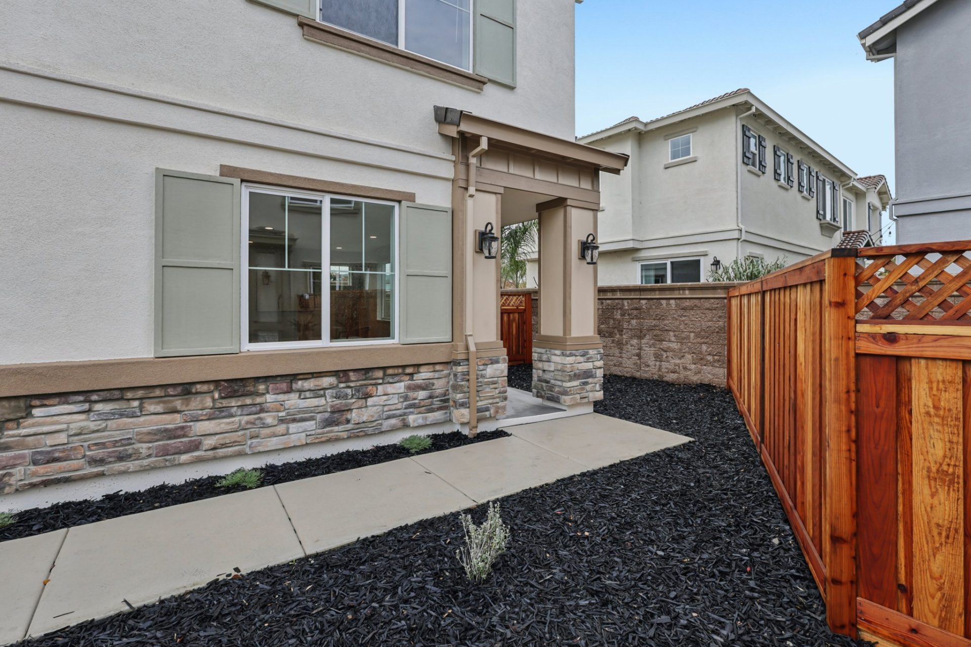 Side yard and front entry of a home with cream stucco, stone veneer wainscoting, and sage green window shutters. Side yard and front entry of a home with cream stucco, stone veneer wainscoting, and sage green window shutters. The covered entry has a wood 