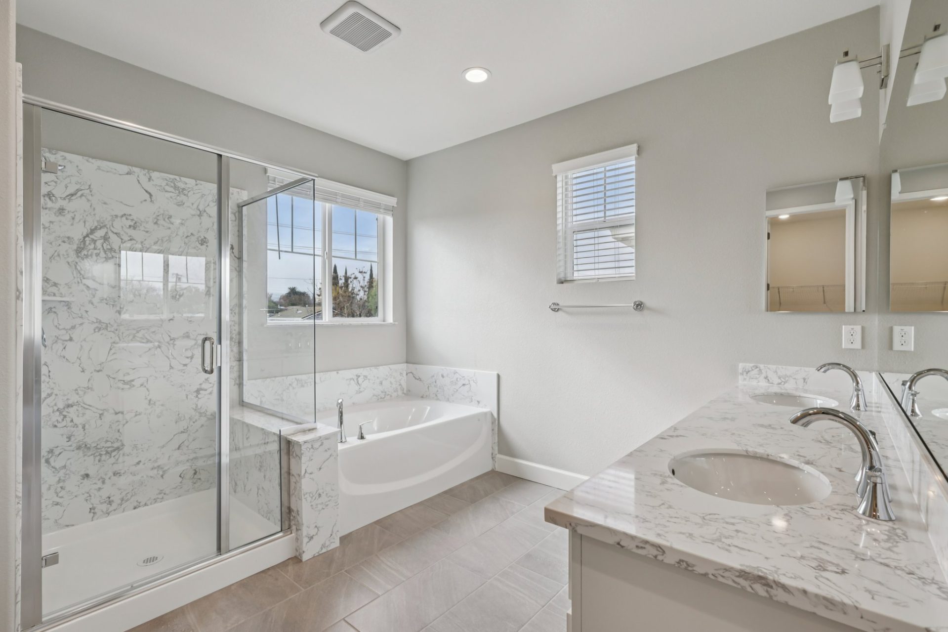 Bathroom with a glass-enclosed shower, a built-in soaking tub, and a dual-sink vanity with marble-look countertops. The space has light gray walls, tile flooring, and a window with white blinds bringing in natural light. The shower features marble-look ti