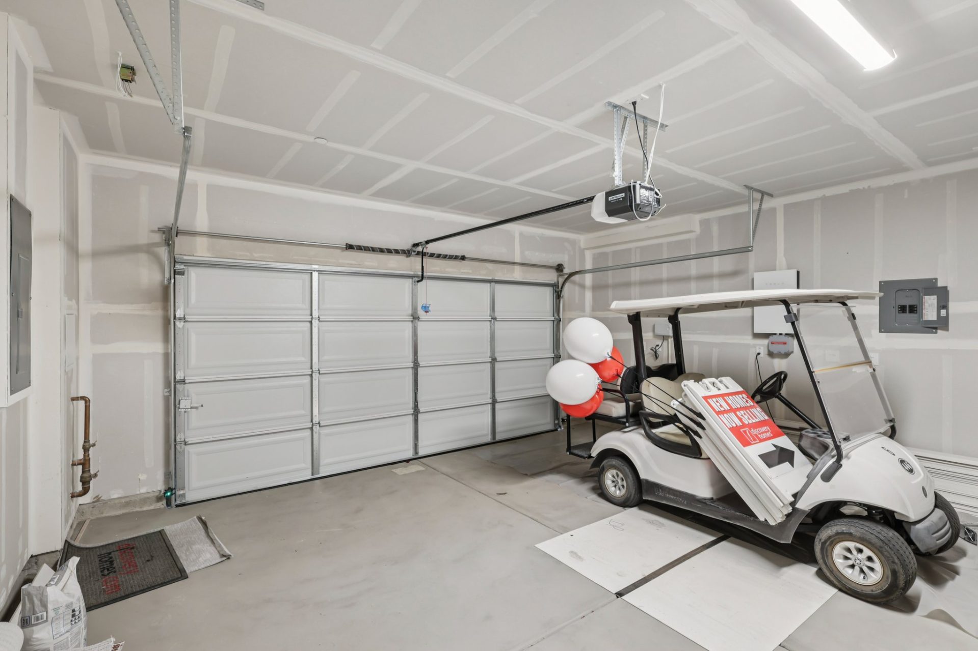 Interior of a finished garage with white walls, a sectional roll-up door, and an automatic garage door opener. A white golf cart with promotional balloons and a sign is parked inside, and the concrete floor shows some markings. The space is well-lit with 