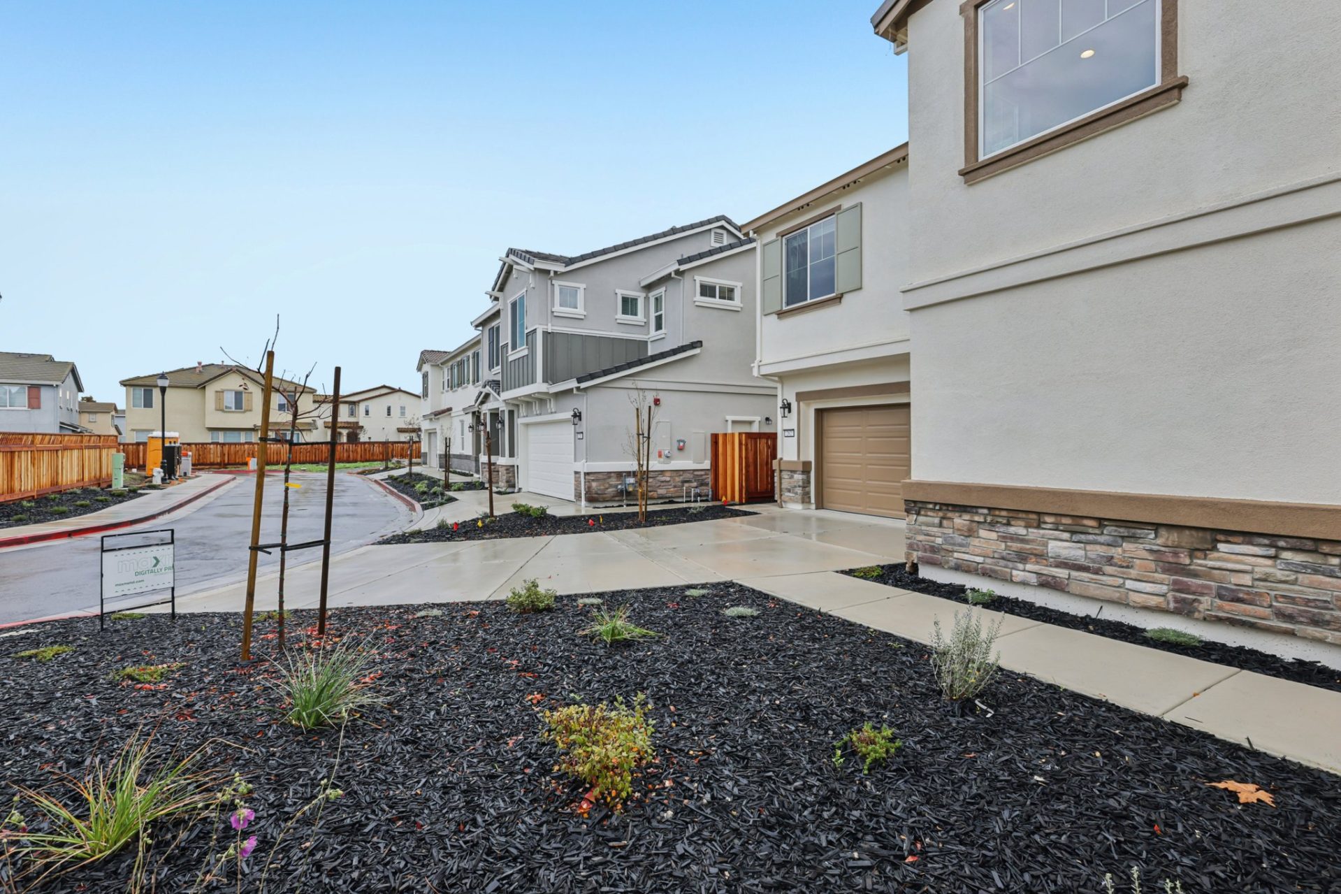 Streetscape of a residential community with two-story homes in cream and tan stucco with tile roofs. The view shows garages, driveways, and young landscaping with black mulch beds and newly planted trees along a wet street. A wood fence and stone veneer a