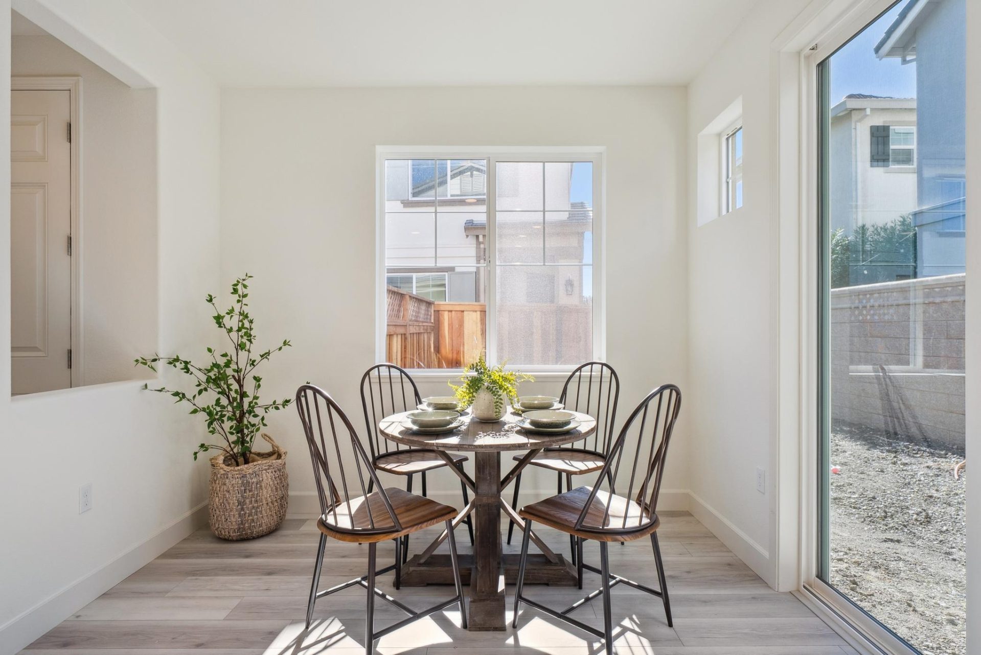 Breakfast nook with a round wood dining table and metal Windsor-style chairs, set beside a window and a sliding glass door. The space has white walls, wood-look flooring, and a potted plant in the corner, with natural light streaming in from the backyard 