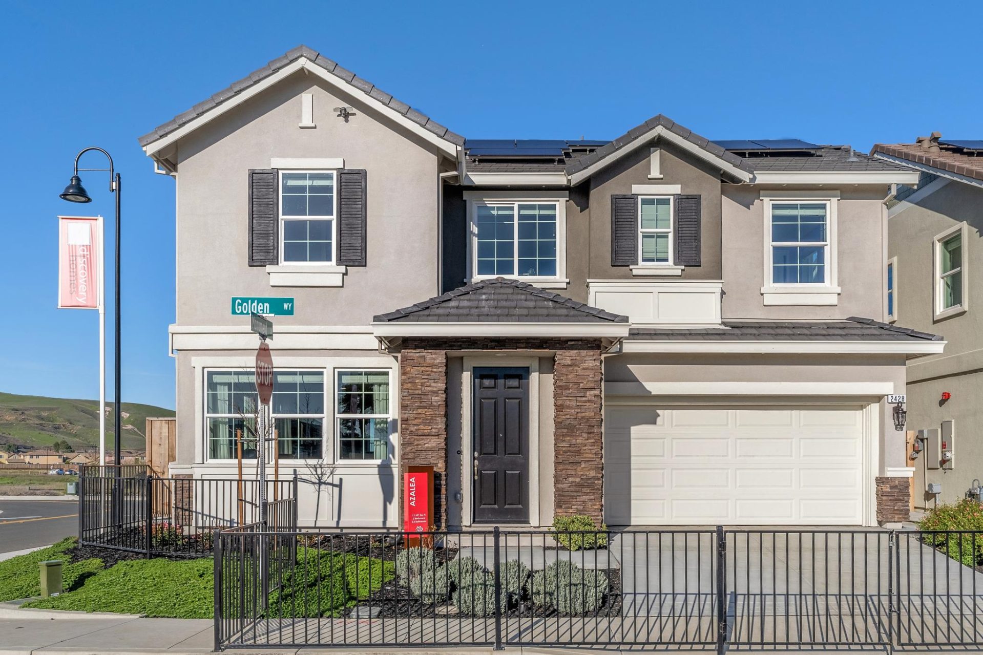 Two-story home with two-tone stucco in cream and tan, featuring decorative window shutters, stone veneer accents, and rooftop solar panels. The home has a two-car garage with a white door, a covered front entry, and a metal fence enclosing the front yard.