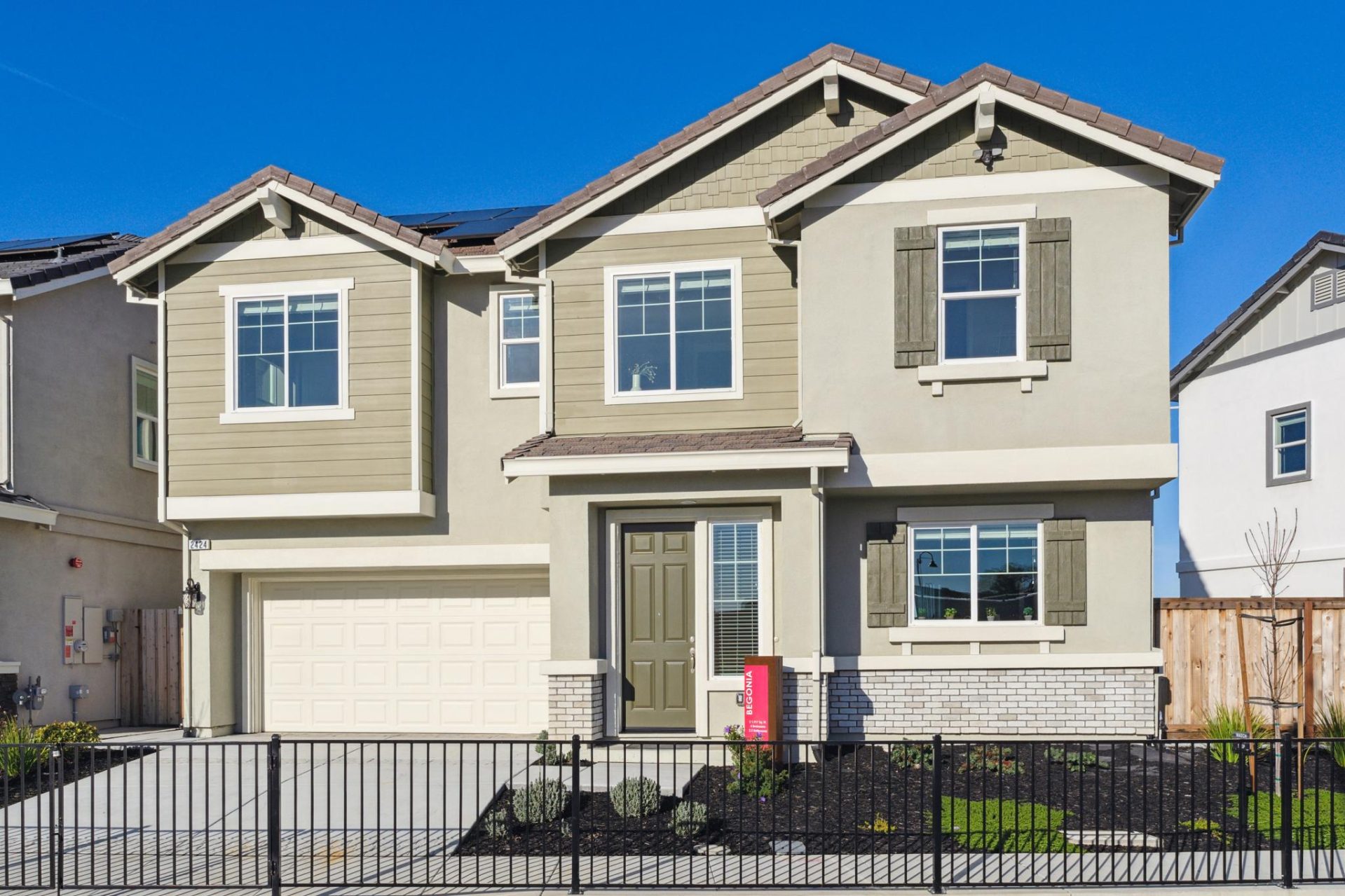 Two-story home with two-tone exterior in tan and sage green, featuring decorative window shutters, mixed siding textures, and a gabled roofline. The home has a two-car garage with a white door, a covered front entry with a green door, and a metal fence wi