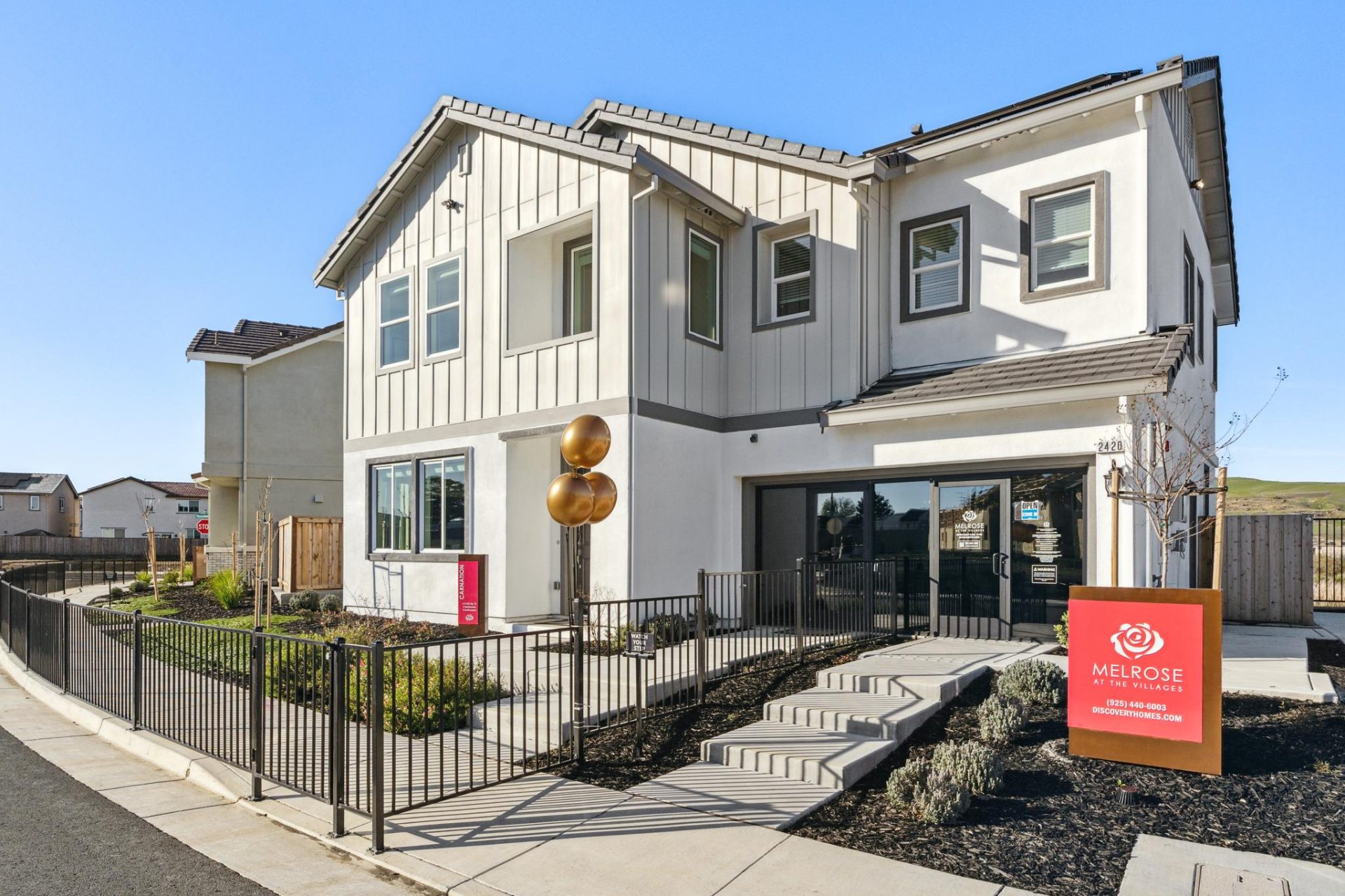 Two-story home with white board-and-batten siding, contrasting darker trim, and a mix of gabled rooflines. The home features large windows, a metal fence enclosing the yard, and a concrete walkway leading to the entry. A "Melrose" sign stands in front, wi