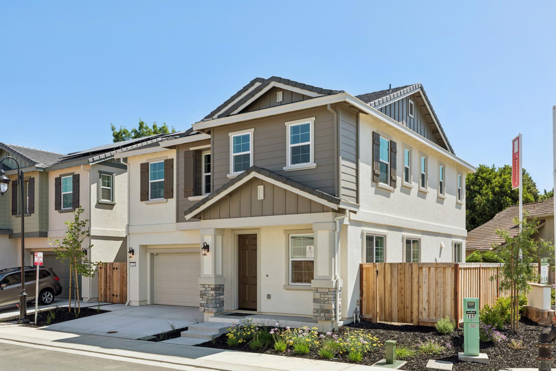 Two-story home with two-tone exterior in taupe and cream, featuring decorative window shutters, stone veneer accents, and a craftsman-style covered entry. The home has a two-car garage with a white door, mixed siding textures, and young landscaping along