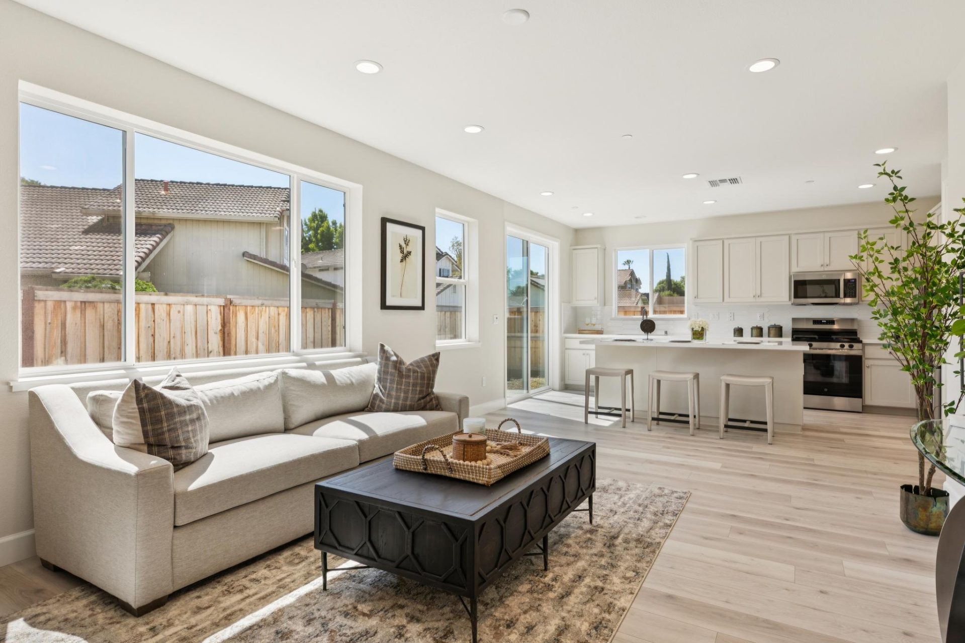 Open-concept living room and kitchen with light walls and wood-look flooring. The living area features a neutral sectional sofa with throw pillows, a dark metal-framed coffee table, and a potted tree, set on a patterned rug. The kitchen beyond has white c