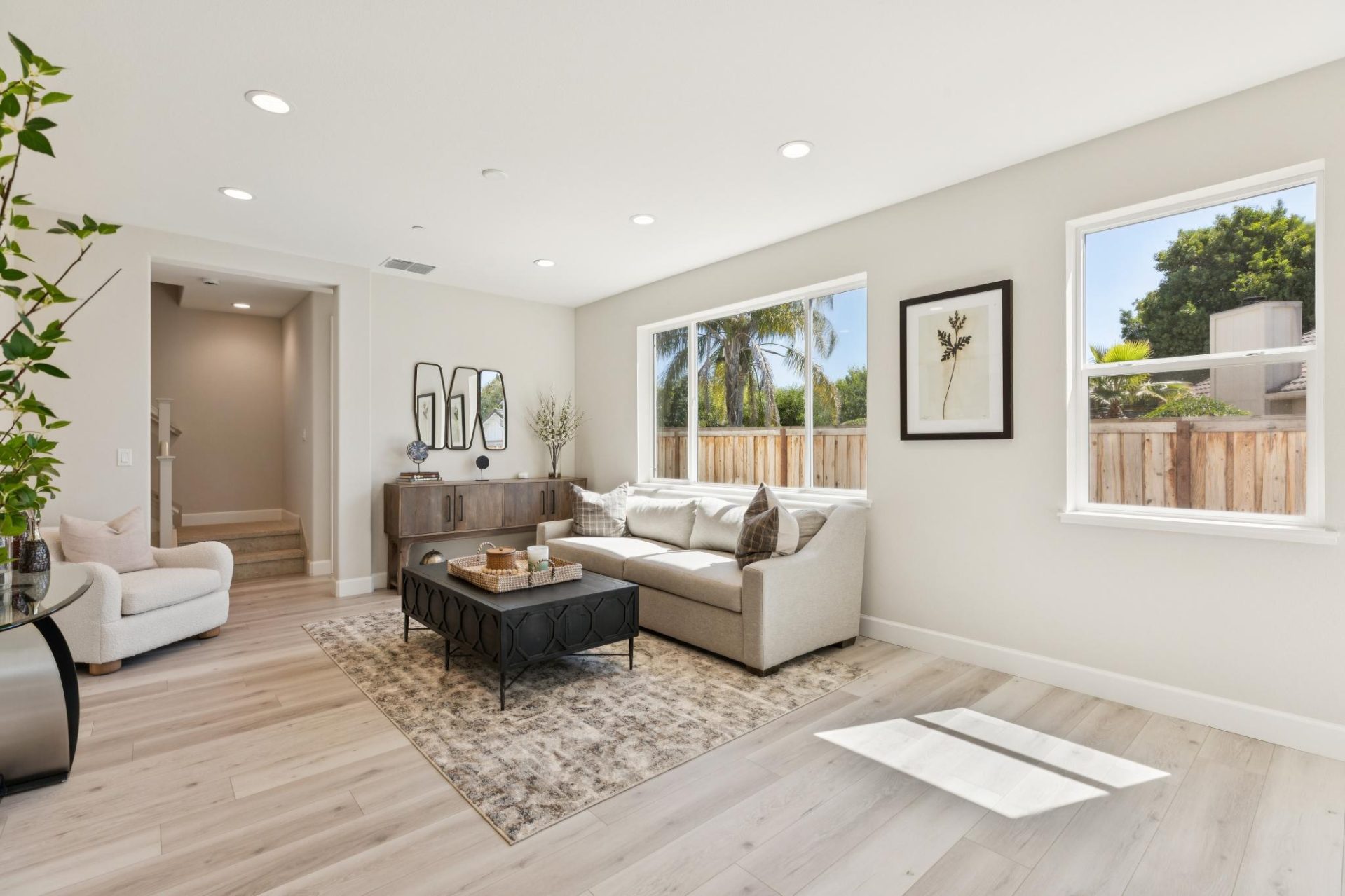 Living room with light gray walls, wood-look flooring, and multiple windows bringing in natural light. The space features a neutral sectional sofa with throw pillows, an accent chair, and a dark upholstered ottoman on a patterned rug, with a wood console