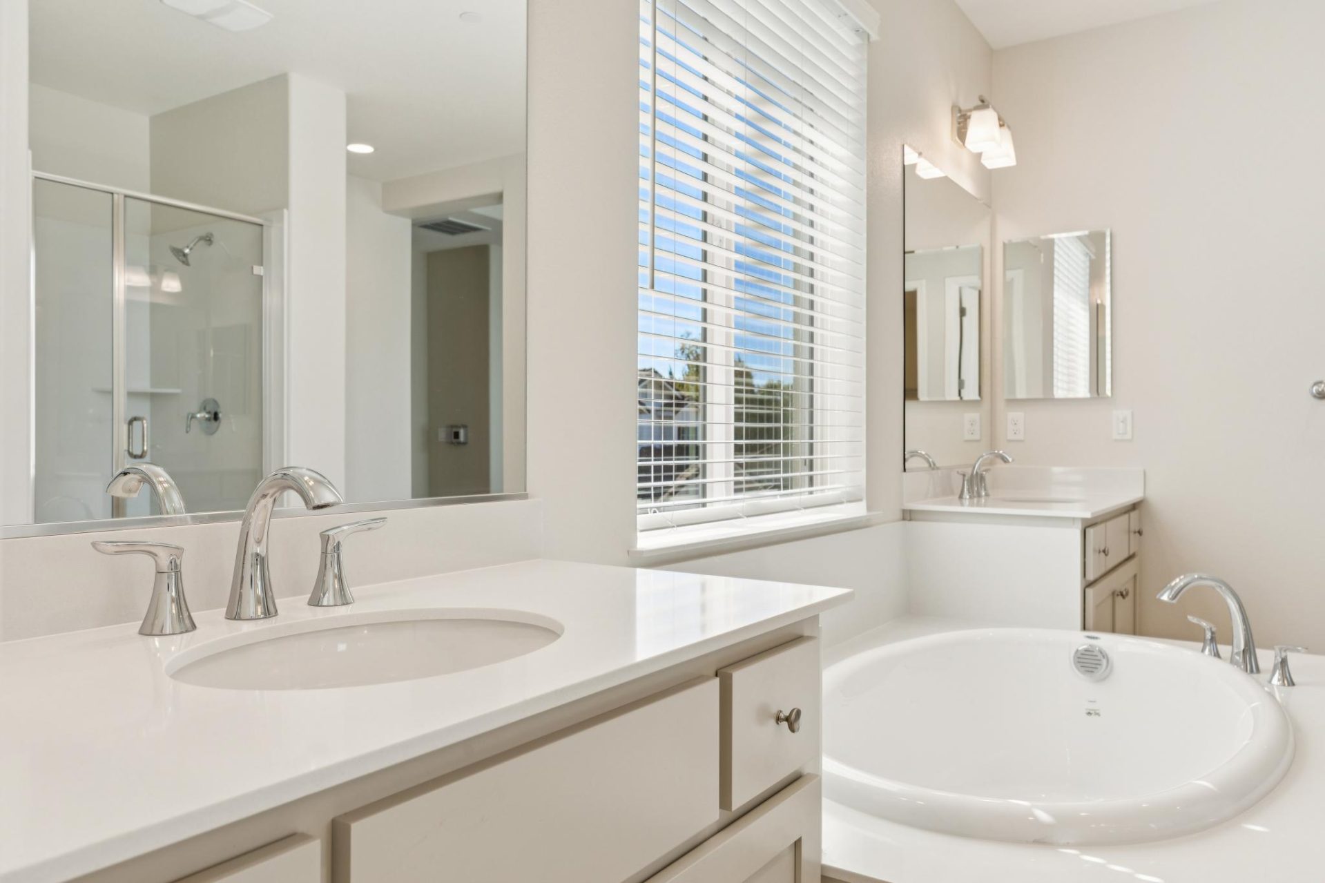 Bathroom with dual vanities, white cabinets, and light countertops, featuring a built-in soaking tub beneath a window with white blinds. The space has large mirrors, sconce lighting, and a glass-enclosed shower, with natural light coming in from the windo