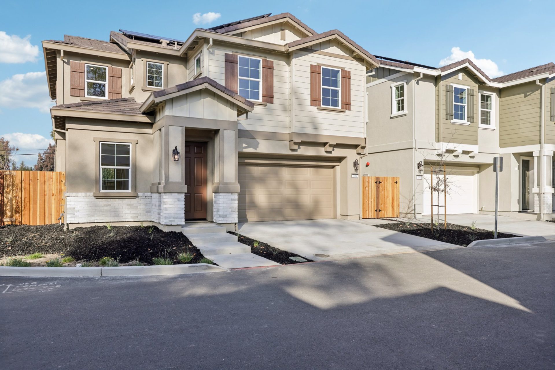 Two-story homes in a residential community, featuring two-tone stucco in taupe and cream with decorative shutters and stone veneer accents. The foreground home has a two-car garage, a covered entry, and rooftop solar panels, with a shared driveway and you