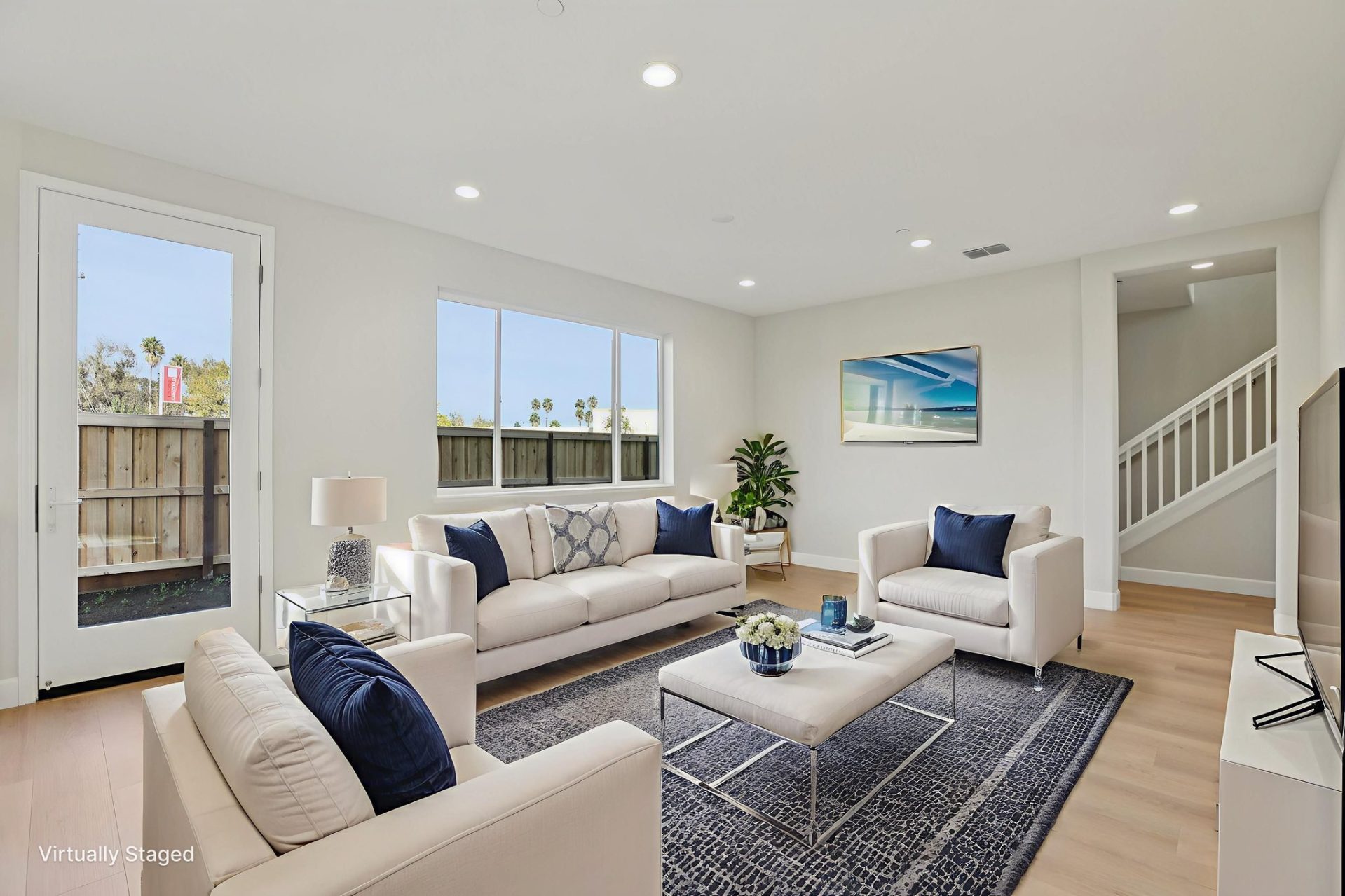 Living room with light walls, wood-look flooring, and natural light from windows and a glass door. The space features two neutral sofas with navy accent pillows, a glass coffee table, and an acrylic side table on a gray rug, with coastal-style wall art. A