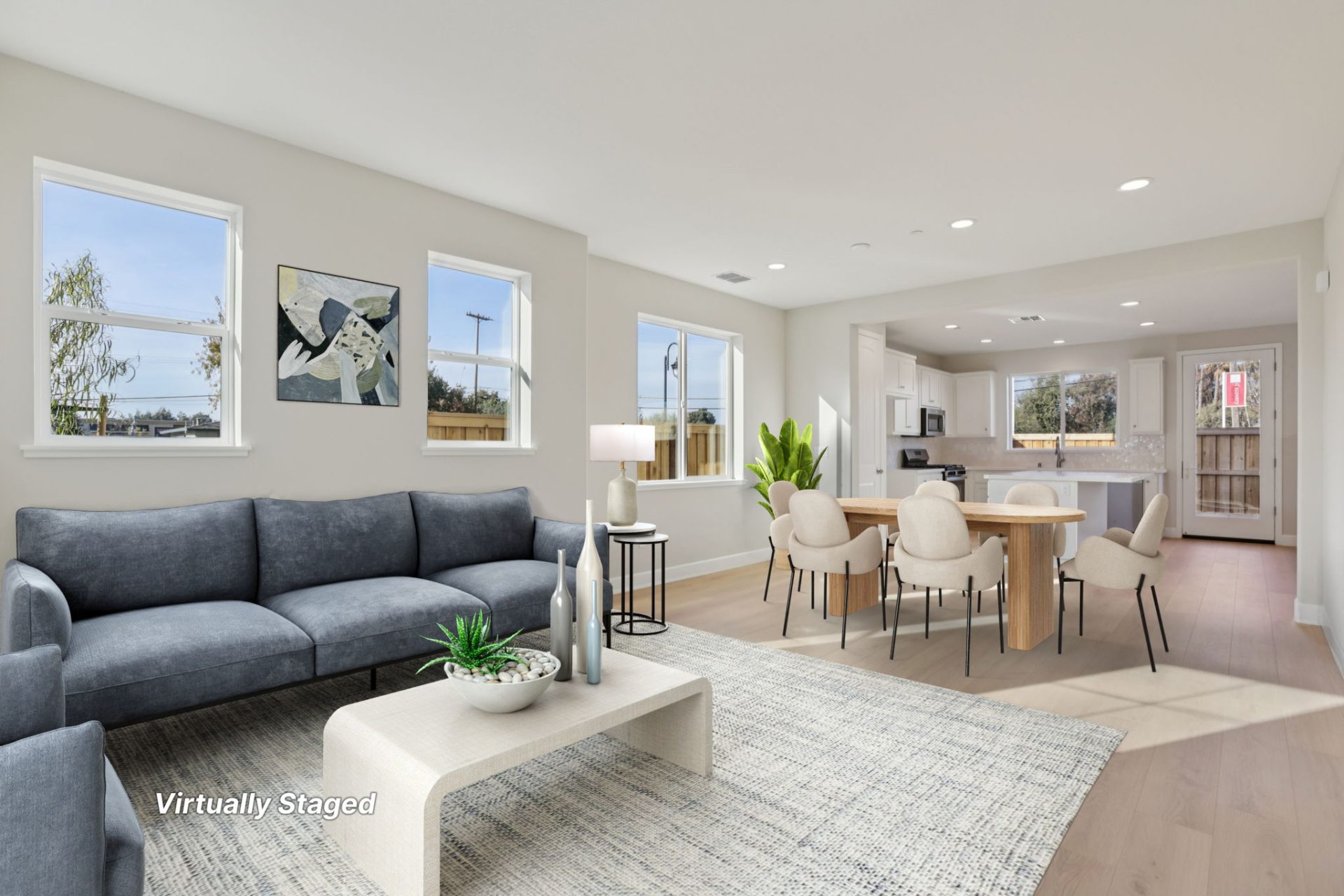 Open-concept living room and kitchen with light walls, wood-look flooring, and multiple windows bringing in natural light. The living area features a gray sofa, a white coffee table, and framed wall art on a patterned rug, flowing into a dining area with 