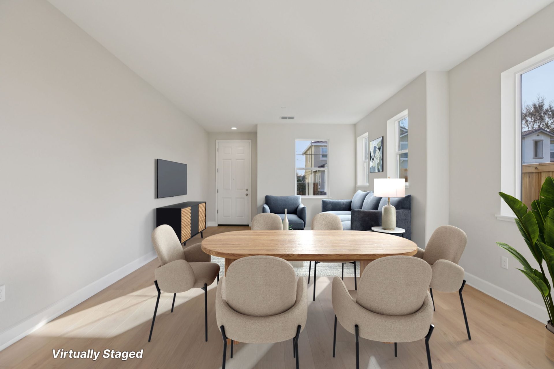 Dining area with a wood table and neutral upholstered chairs, set on light wood-look flooring with white walls. The space connects to a sitting area with accent chairs and a lamp, with a white door and windows bringing in natural light. A potted plant is 