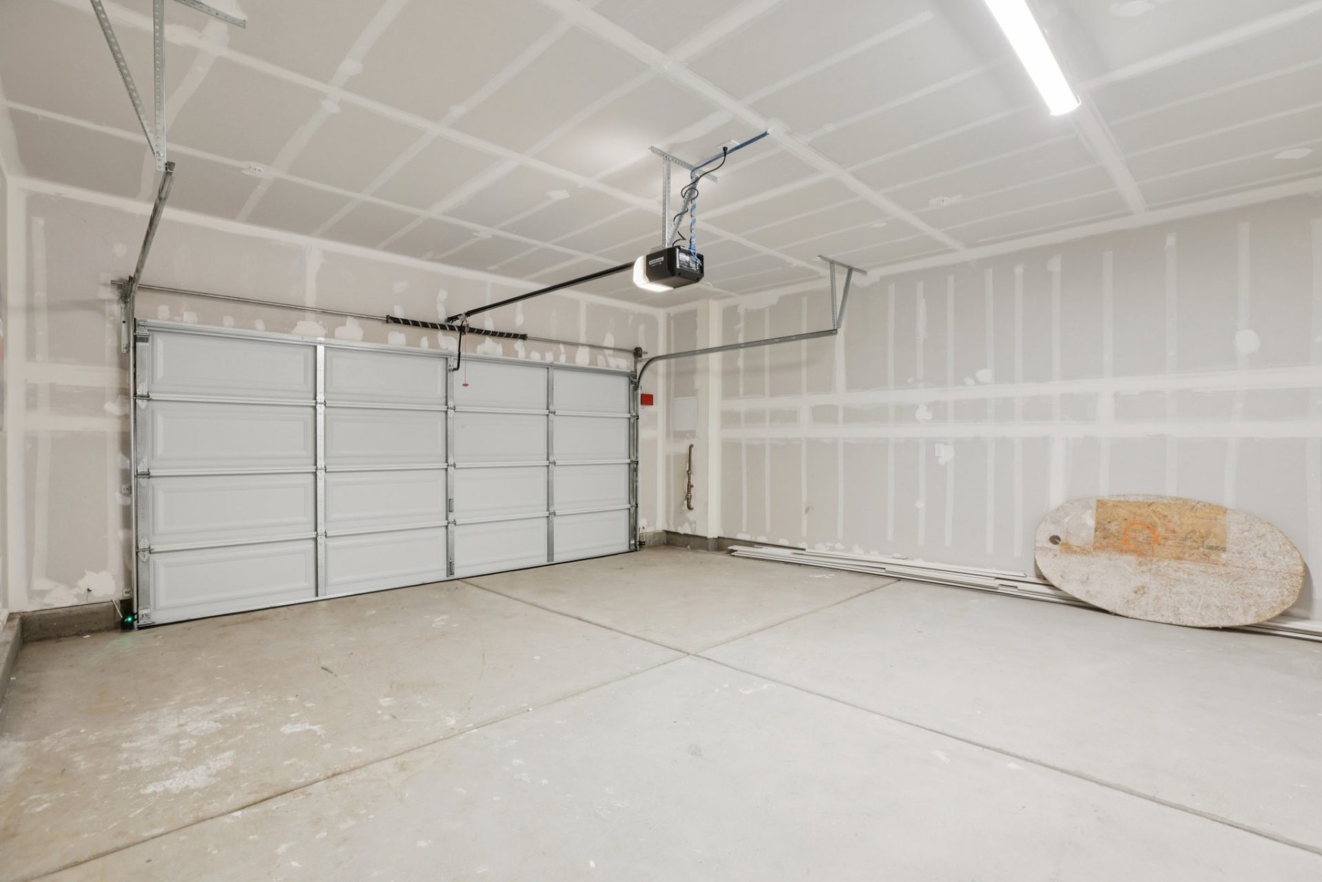 Interior of a finished two-car garage with white walls, a sectional roll-up door, and an automatic garage door opener. The space has a concrete floor, overhead fluorescent lighting, and exposed framing on one wall. A round object leans against the wall on