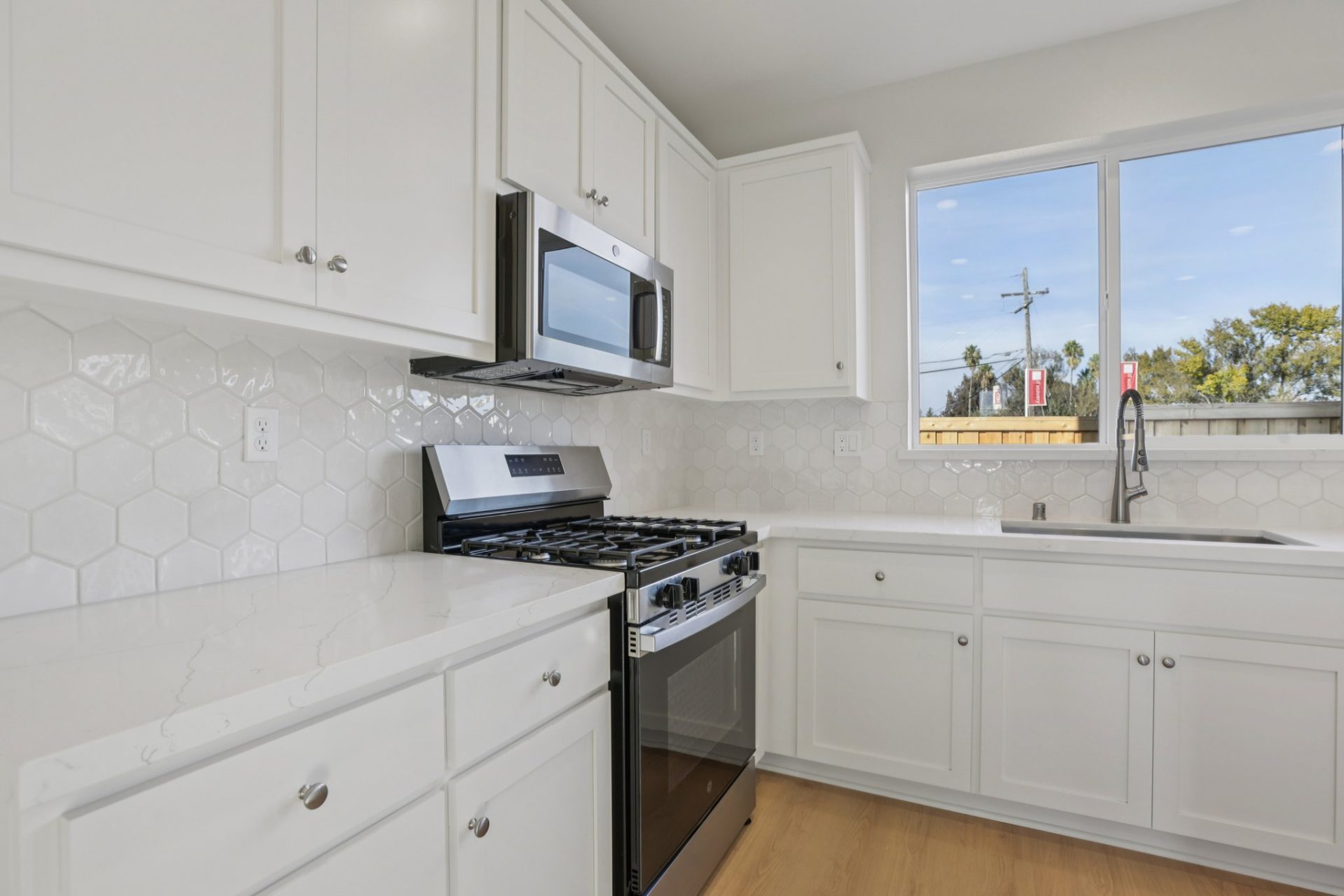 Kitchen with white shaker cabinets, marble-look countertops, and a hexagonal tile backsplash. The space features a stainless steel gas range and over-the-range microwave, with a window above the sink bringing in natural light. The bright, clean finishes g