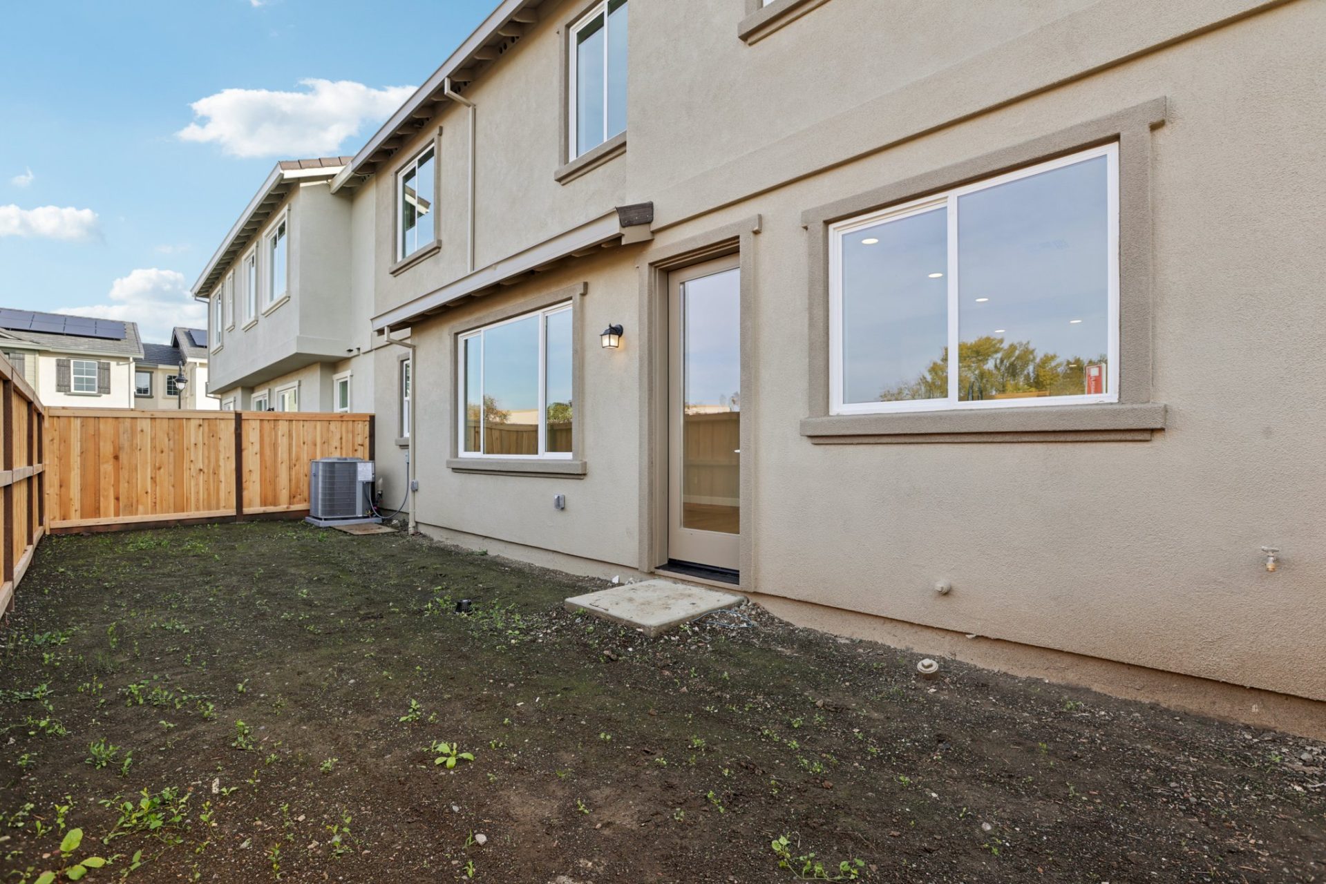 Backyard view of a two-story home with tan stucco, showing a back entry door, windows, and an exterior air conditioning condenser unit. The dirt lot is enclosed by a wood privacy fence, with a neighboring home and rooftop solar panels visible. The space i