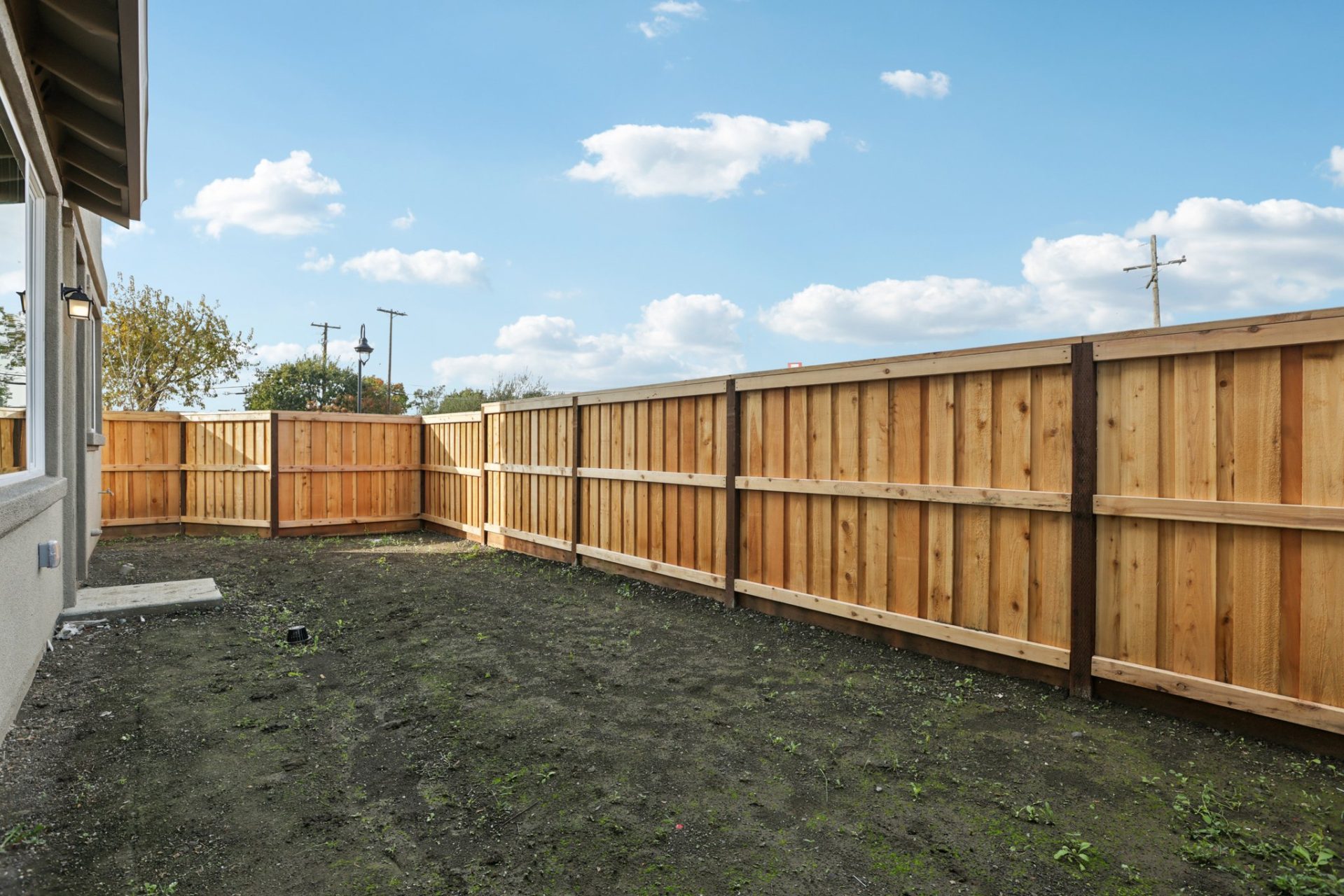 Backyard with a wood privacy fence enclosing a dirt lot ready for landscaping. Backyard with a wood privacy fence enclosing a dirt lot ready for landscaping. The space is bordered by the home on the left, with a side yard visible, under a blue sky with sc