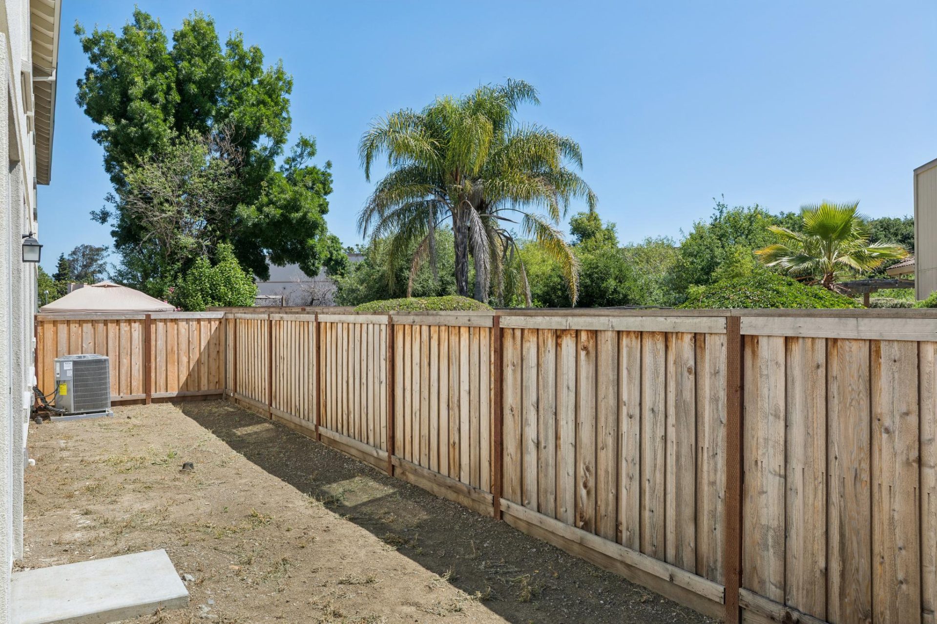 Backyard with a wood privacy fence enclosing a dirt lot ready for landscaping. The space includes an exterior air conditioning condenser unit near the home, with mature palm and shade trees visible beyond the fence under a clear blue sky. The open soil of