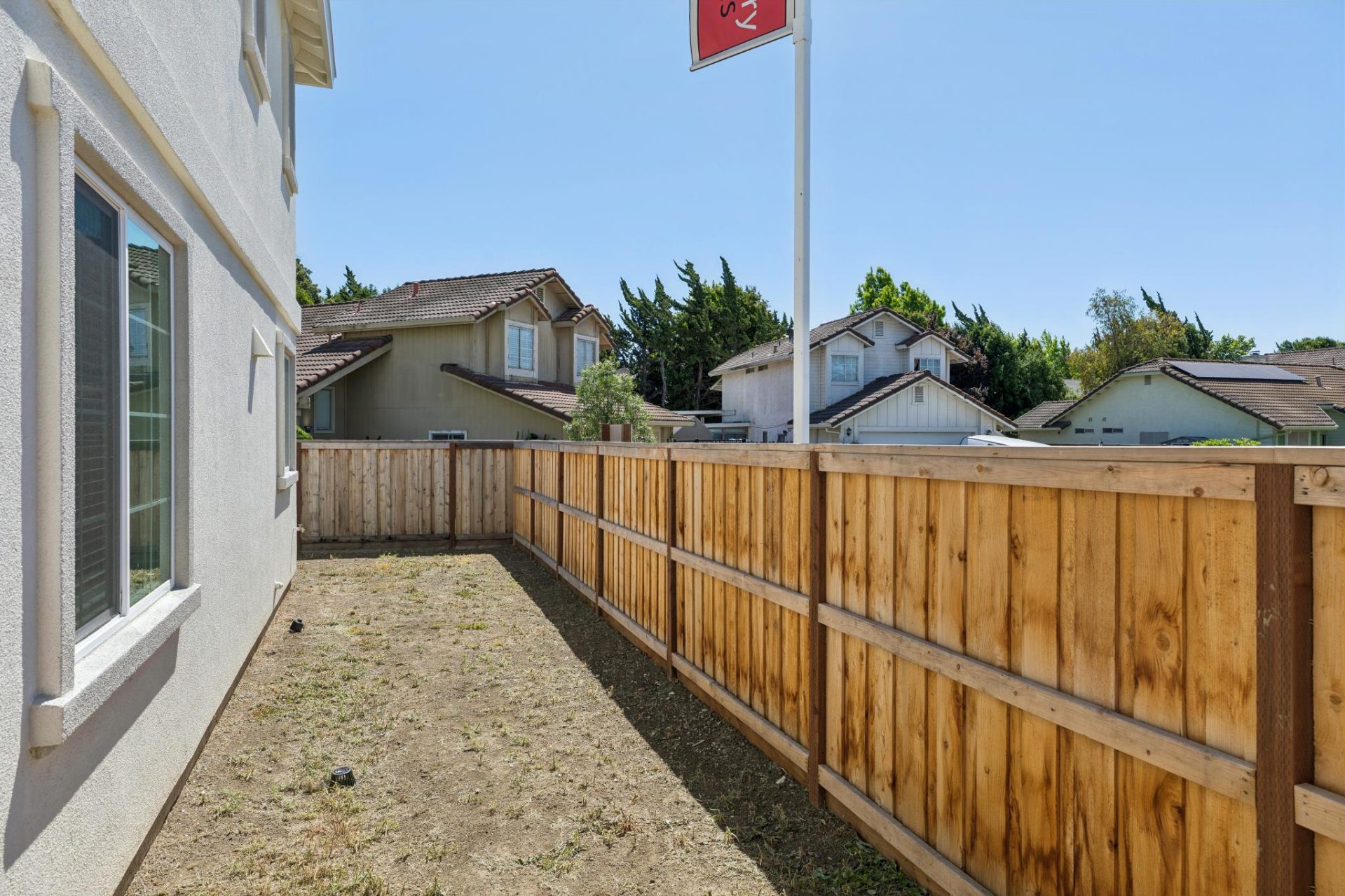 Side yard with a wood privacy fence enclosing a dirt strip alongside the home's tan stucco exterior. The space includes a window and is bordered by neighboring homes visible over the fence, under a clear blue sky. The open soil offers a blank slate for la