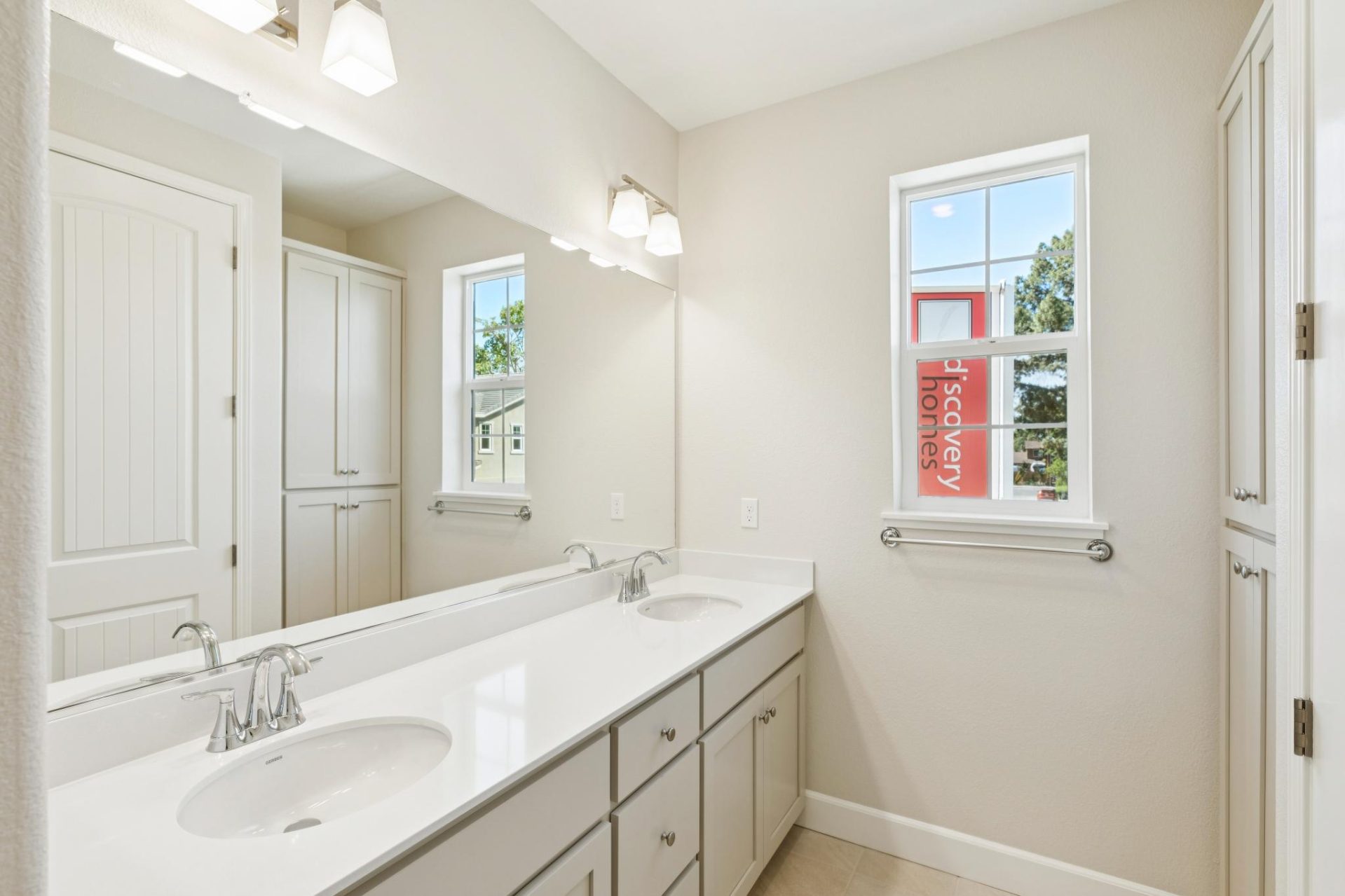 Bathroom with a dual-sink vanity, beige cabinets, and a light countertop, with a large mirror above. A window brings in natural light, and sconce lighting is mounted overhead, with tile flooring and a linen cabinet to the side. The clean, bright finishes 