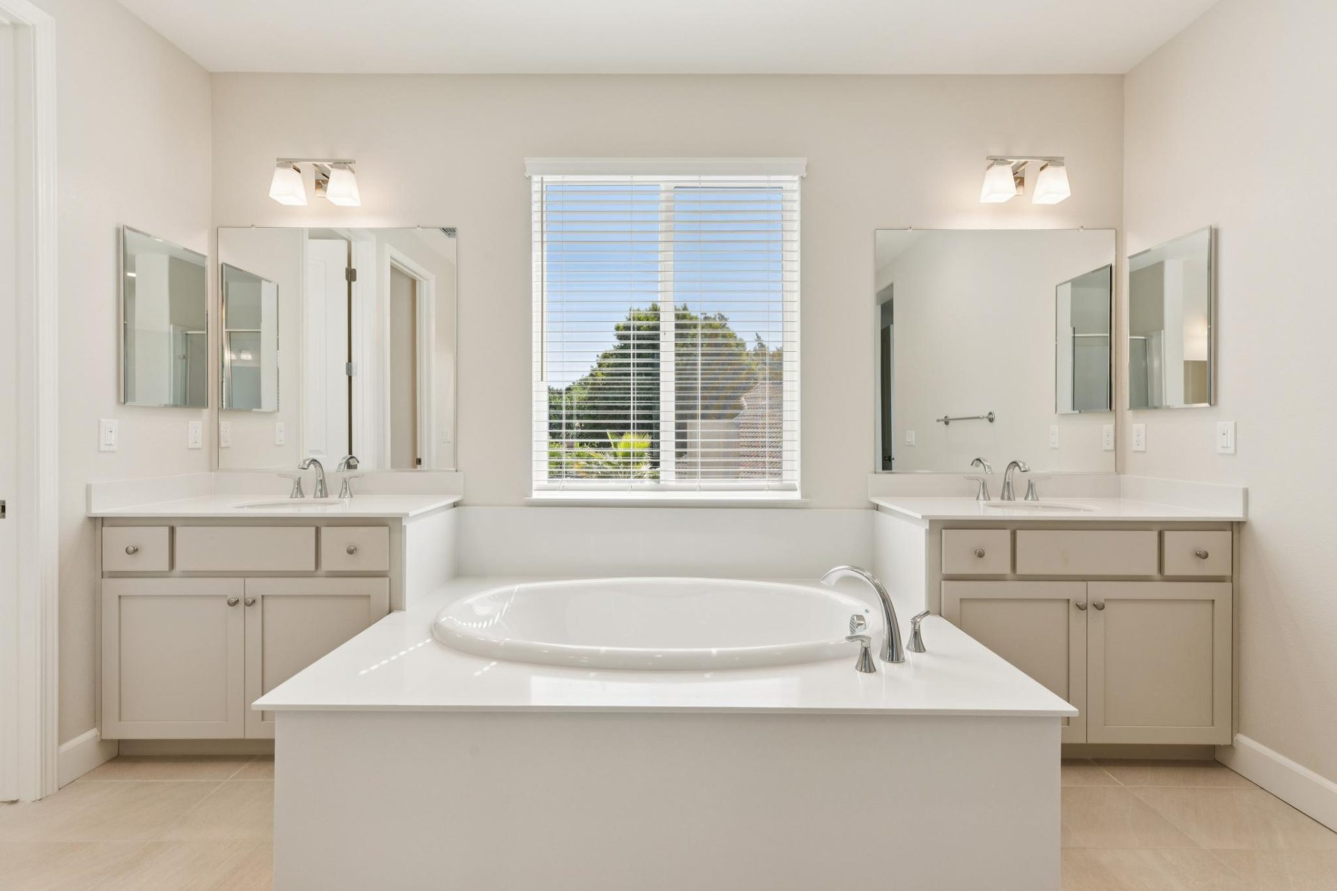 Bathroom with dual vanities, beige cabinets, and light countertops, featuring a built-in soaking tub beneath a window with white blinds. The space has large framed mirrors, sconce lighting, and tile flooring, with natural light coming in from the window. 