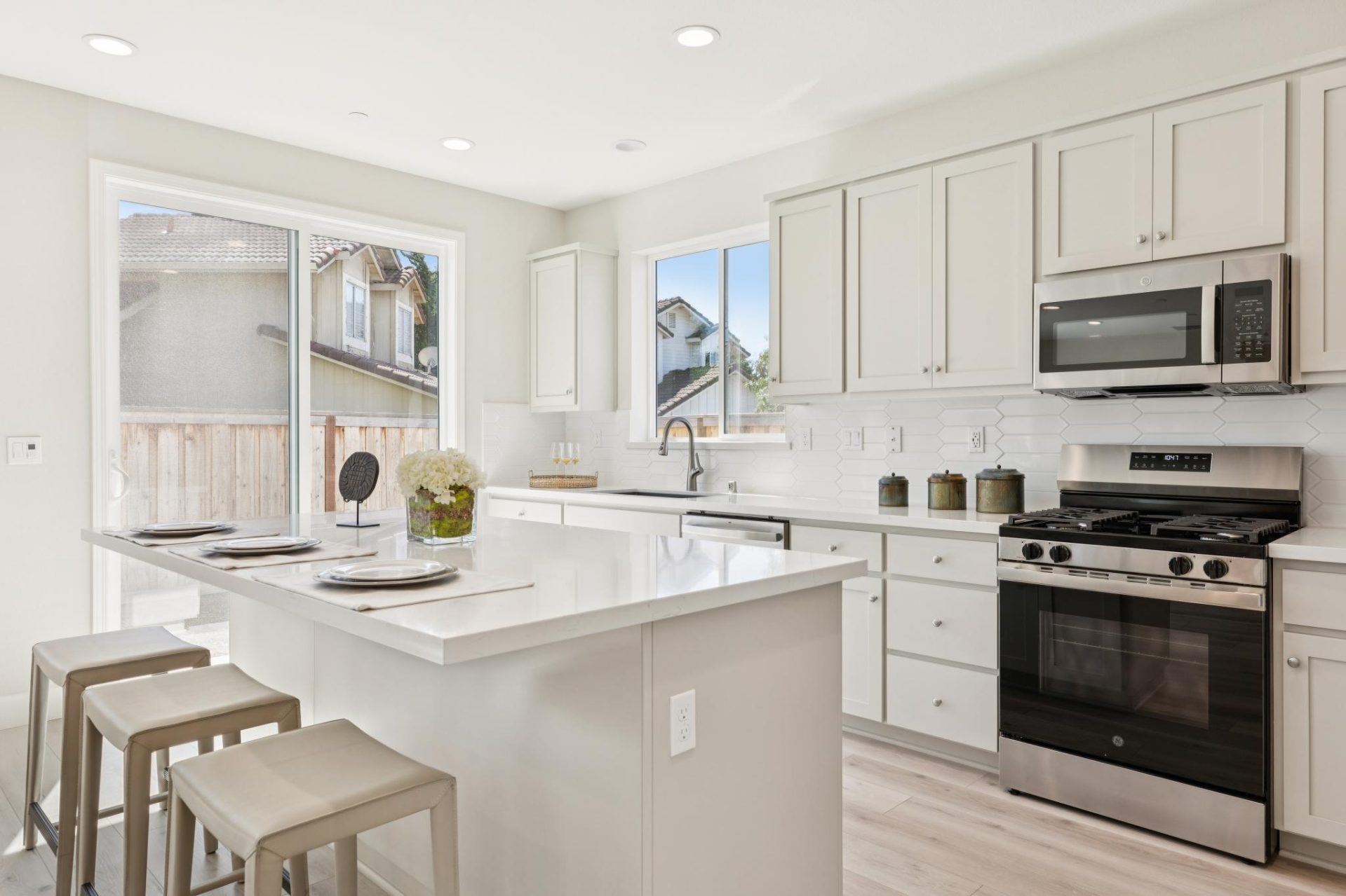 Kitchen with white shaker cabinets, marble-look countertops, and stainless steel appliances. The space features a center island with bar stools, a gas range with microwave above, and a window over the sink bringing in natural light. Light wood flooring an