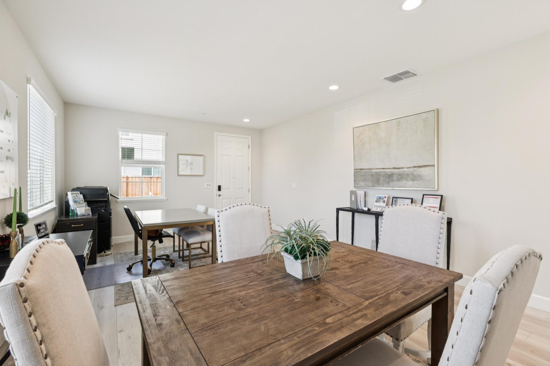 Dining area with a wood table and upholstered chairs, set on wood-look flooring with light walls. The space features a potted plant centerpiece, a home office setup with a desk in the background, and framed wall art. A white front door and a window with b