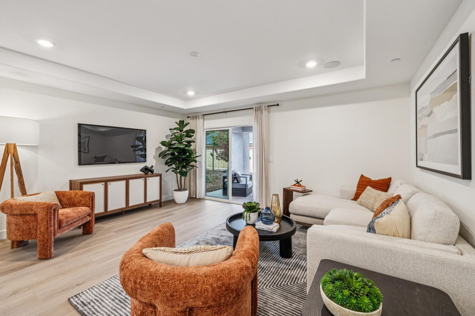 Living room with light walls, wood-look flooring, and a tray ceiling with recessed lighting. Living room with light walls, wood-look flooring, and a tray ceiling with recessed lighting. The space features a neutral sectional sofa, rust-colored accent chai