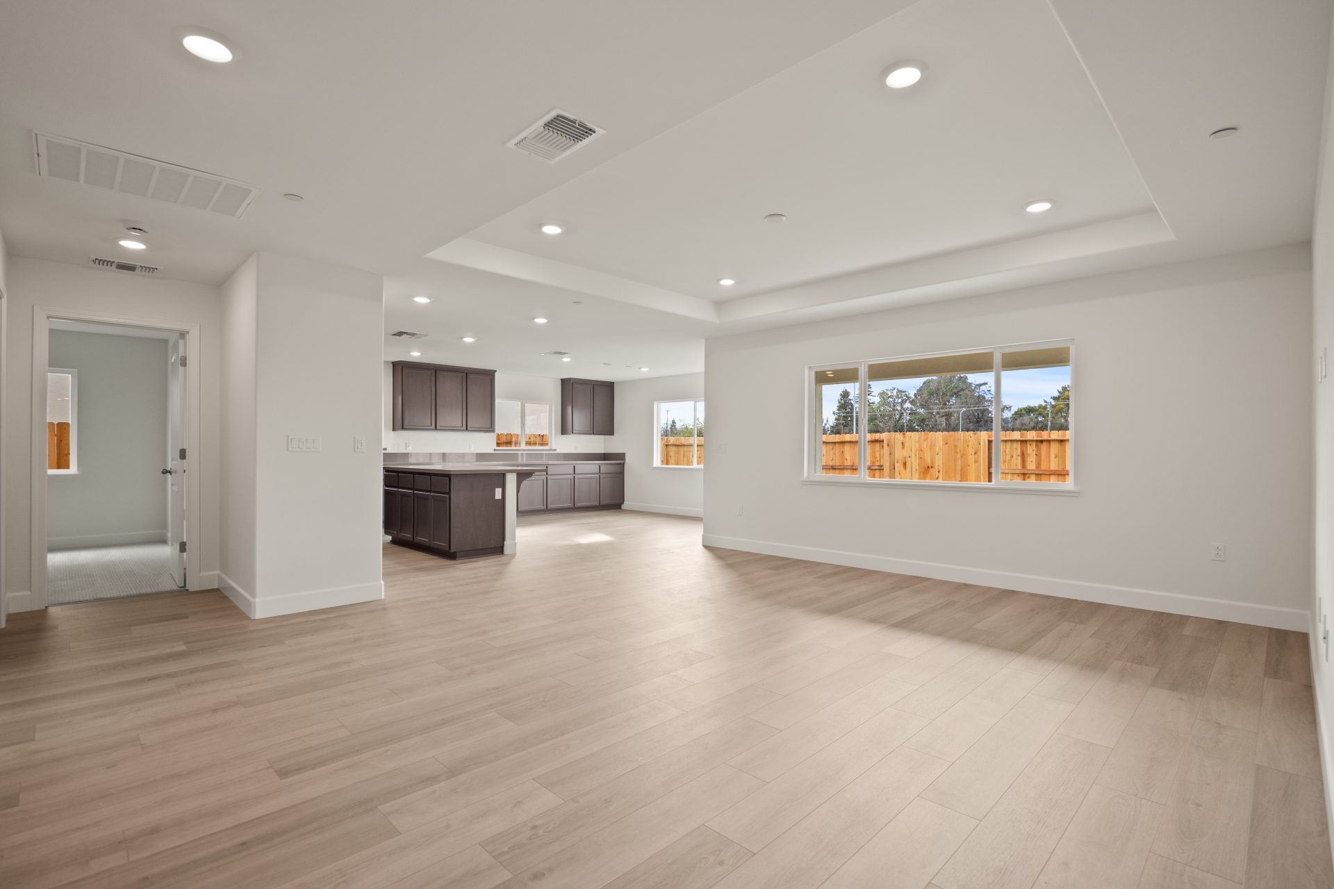 Open-concept main living area with light walls, wood-look flooring, and a tray ceiling with recessed lighting. The kitchen features dark wood cabinets and an island, with a window offering a backyard view of a wood fence. The empty, bright space showcases