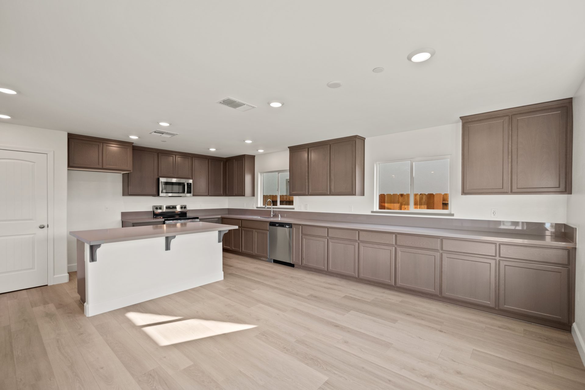 Kitchen with dark wood cabinets, light countertops, and stainless steel appliances. The space features a center island, a gas range with microwave above, and a window above the sink bringing in natural light. Light wood flooring and recessed lighting comp