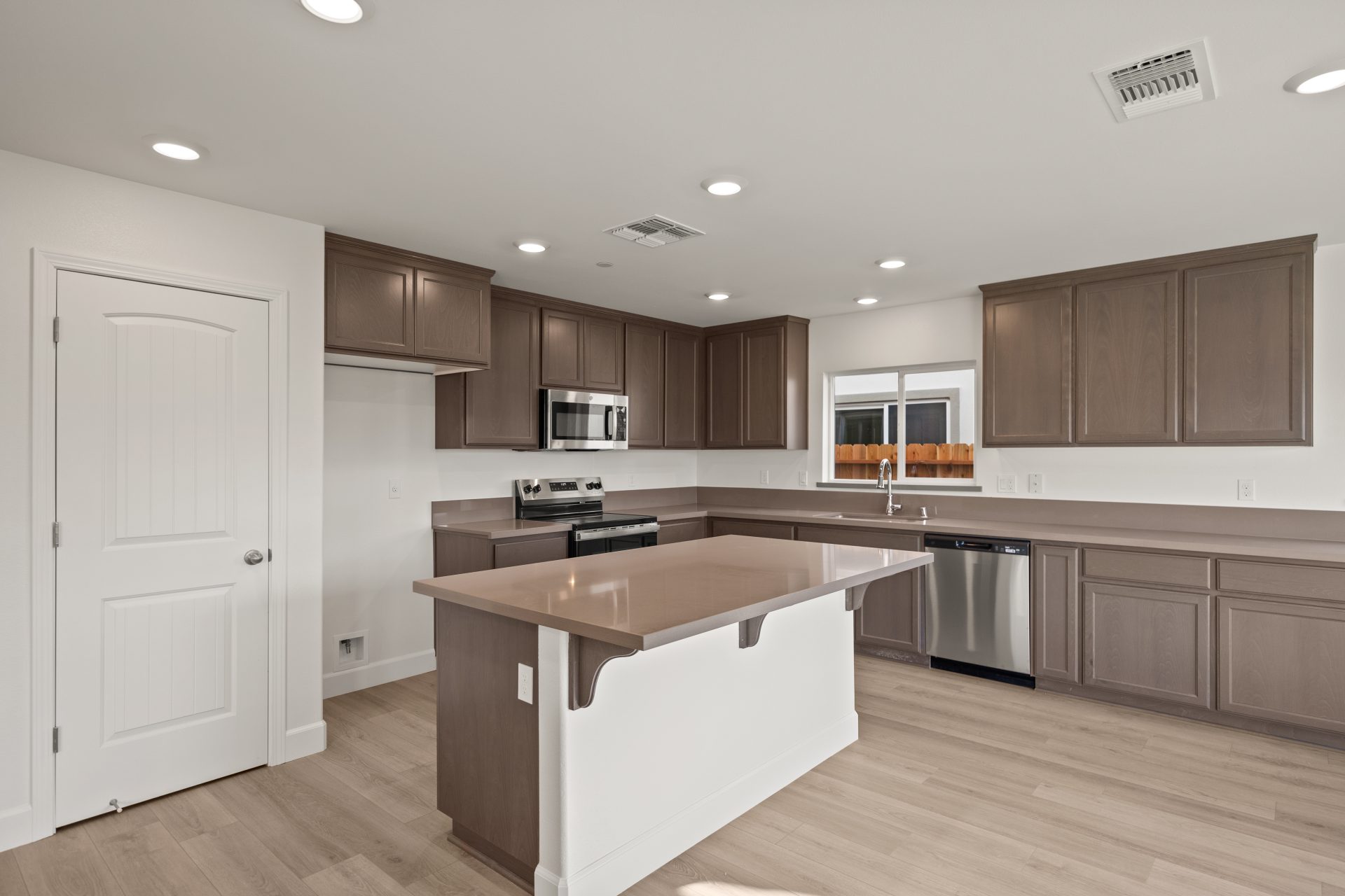 Kitchen with dark wood cabinets, light countertops, and stainless steel appliances. The space features a center island with a white base, a gas range with microwave above, and a window over the sink bringing in natural light. A white pantry door, light wo