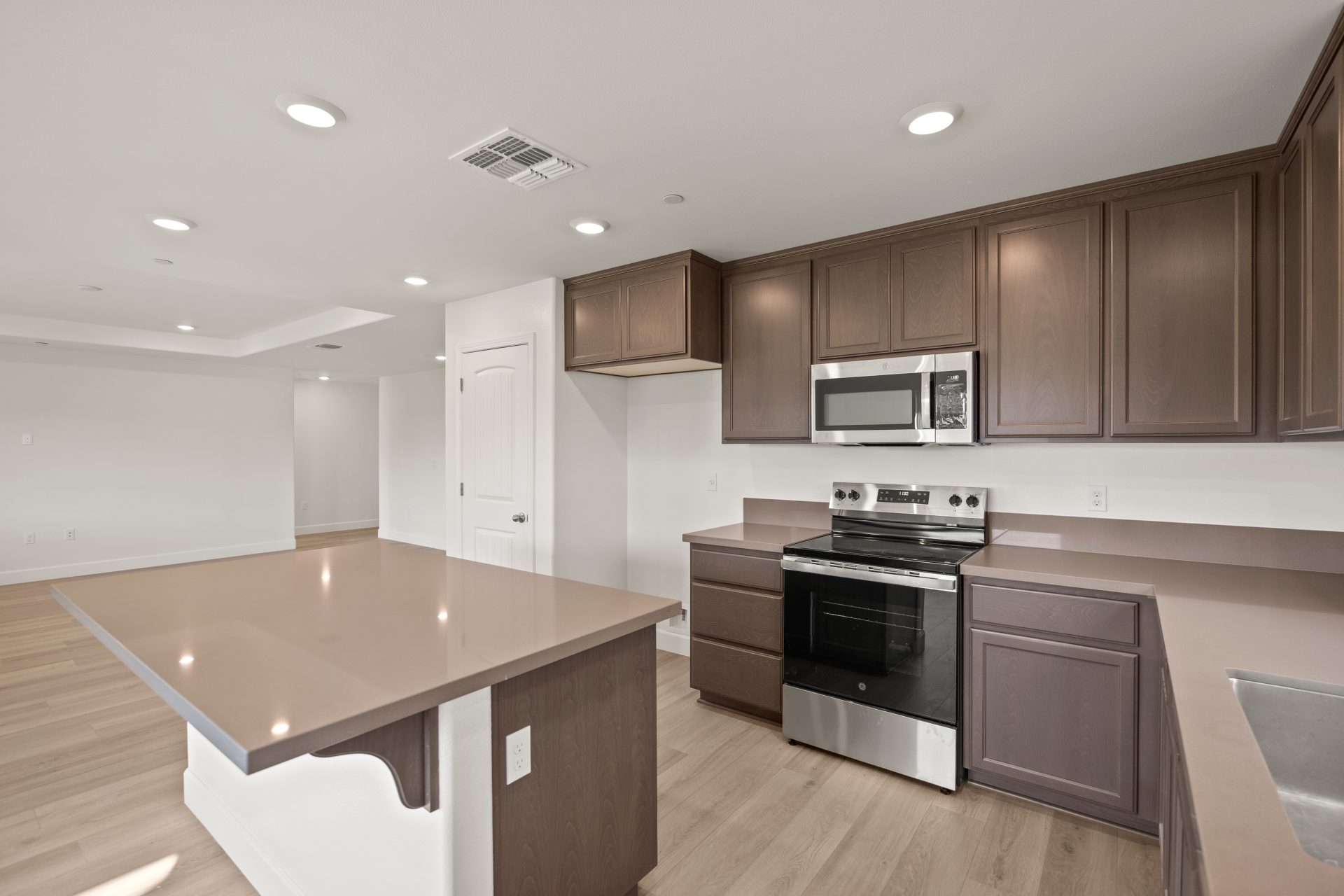 Kitchen with dark wood cabinets, light countertops, and stainless steel appliances including an electric range and over-the-range microwave. The space features a center island with a white base, light wood flooring, and recessed lighting. The open layout 