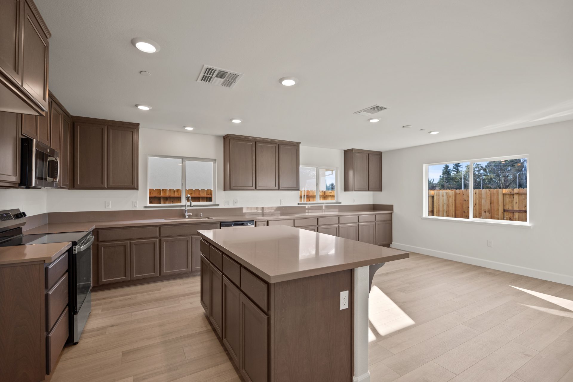 Open-concept living area with light walls, wood-look flooring, and recessed lighting. A kitchen island with dark wood cabinets and a light countertop anchors the space, with multiple windows offering backyard views of a wood fence. The empty, bright room 