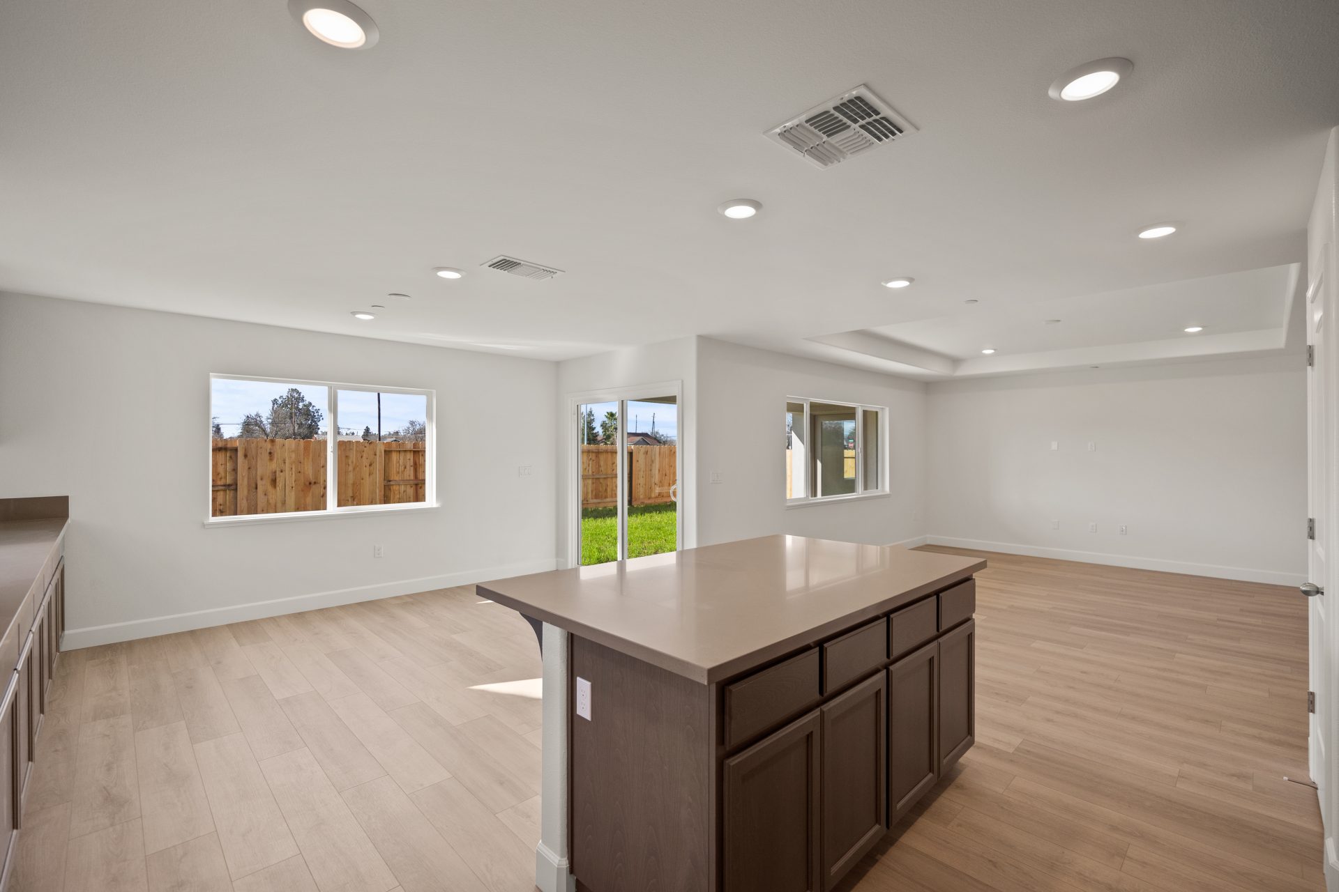 Open-concept living area with light walls, wood-look flooring, and recessed lighting. A kitchen island with dark wood cabinets and a light countertop anchors the space, with multiple windows offering backyard views of a wood fence. The empty, bright room 