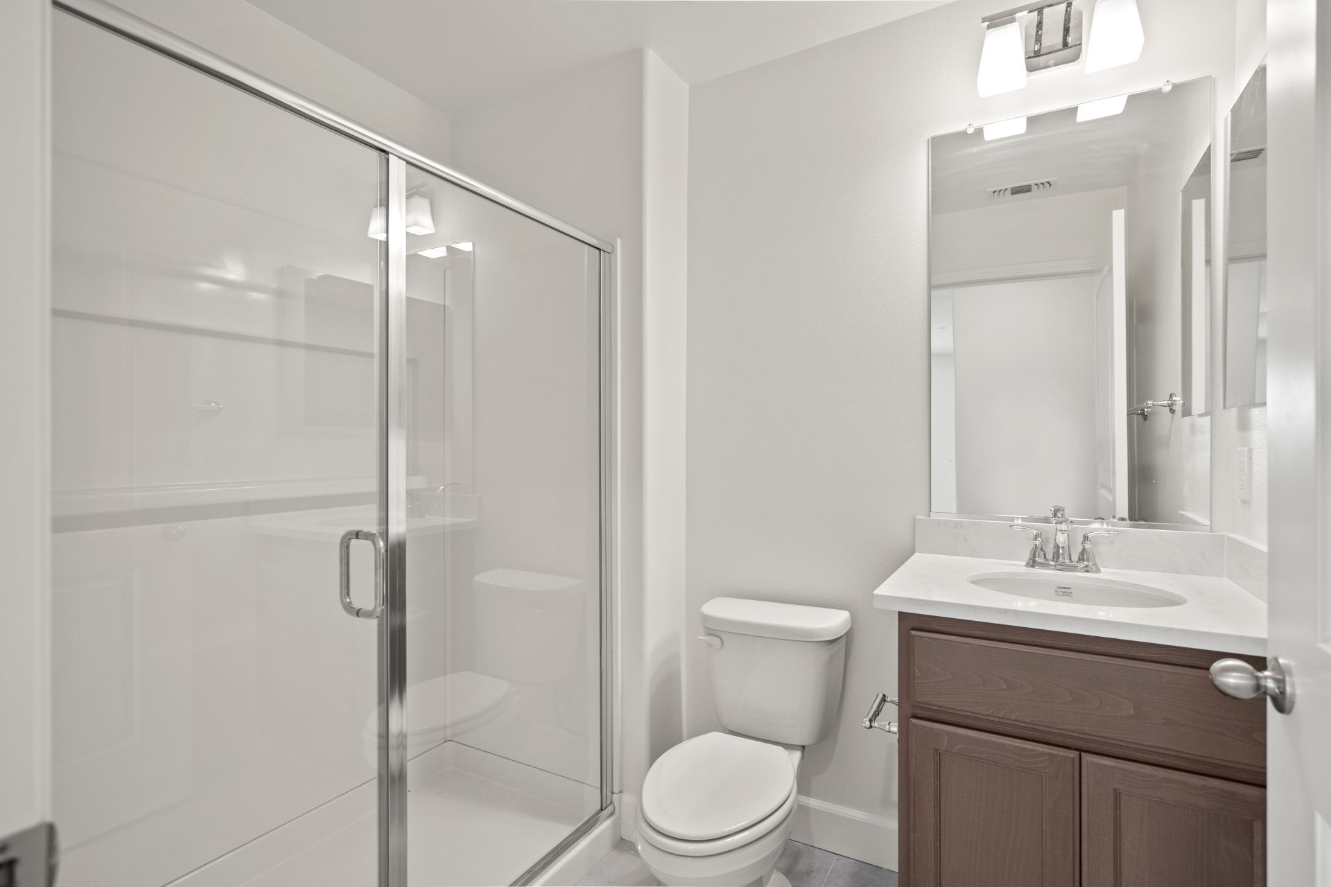 Bathroom with a glass-enclosed shower, a single-sink vanity with dark wood cabinets, and a light countertop. The space has white walls, a large mirror, and sconce lighting above, with tile flooring. The clean, bright finishes give the room a modern, funct