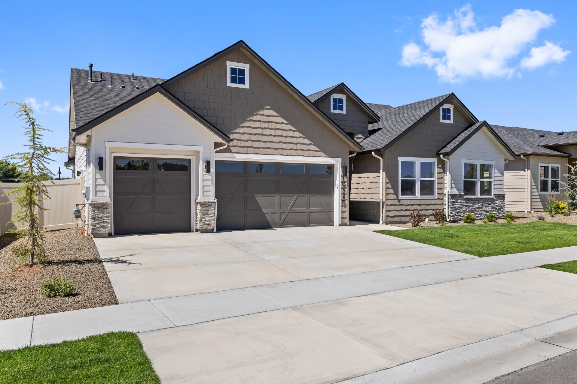 Single-story home with two-tone exterior in taupe and tan, featuring mixed siding textures, stone veneer accents, and a craftsman-style design. The home has a three-car tandem garage with two doors, gabled rooflines, and young landscaping along a wide con
