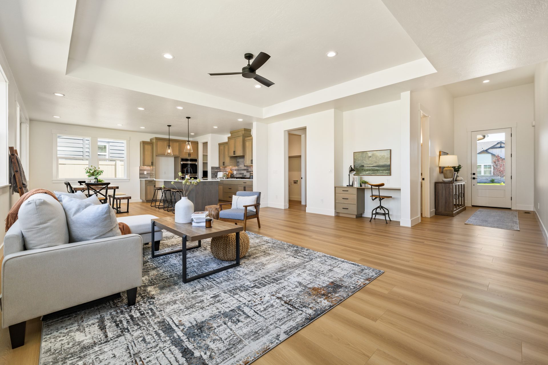 Open-concept living area with wood flooring, light walls, and a tray ceiling with a ceiling fan. The living room features a neutral sofa, accent chairs, and coffee tables on a patterned rug, flowing into a kitchen with an island and a dining nook beyond.