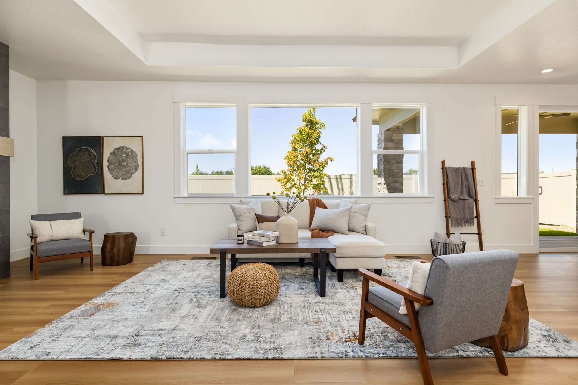 Living room with wood flooring, light walls, and a tray ceiling, with large windows bringing in natural light. The space features a neutral sofa, accent chairs, a coffee table, and a woven pouf on a patterned rug, with framed art and a decorative ladder.