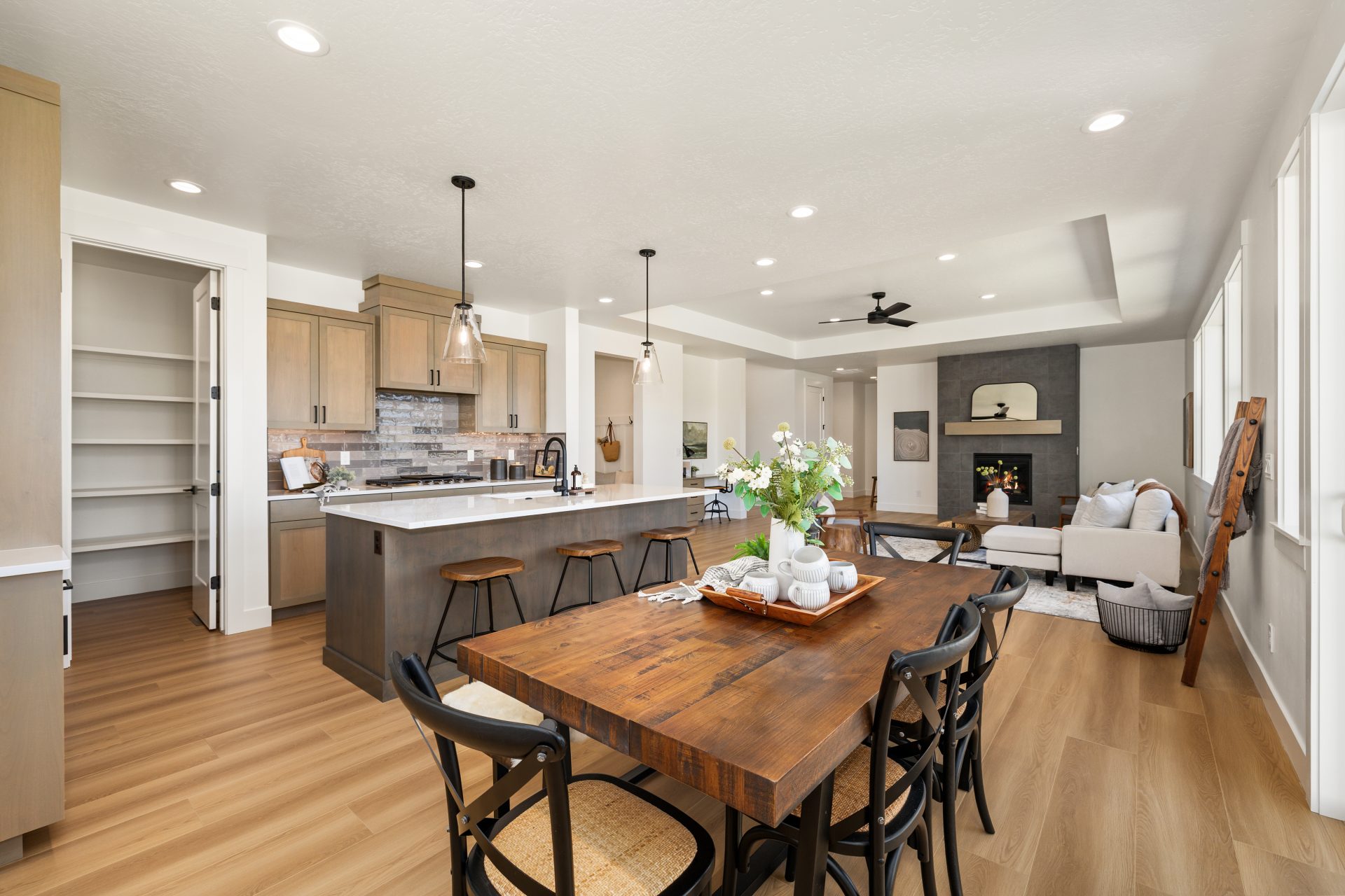 Open-concept kitchen, dining, and living area with wood flooring and light walls. The kitchen features wood cabinets, an island with bar stools and pendant lighting, and a walk-in pantry, with a wood dining table in the foreground. The living area beyond
