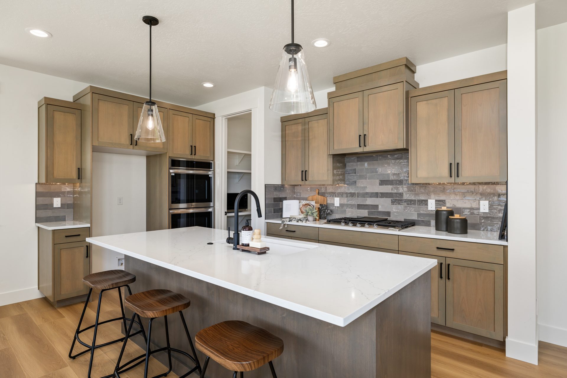 Kitchen with wood cabinets, marble-look countertops, and a tile backsplash. The center island features pendant lighting, a waterfall-edge countertop, and bar stools, with stainless steel appliances including a built-in oven and gas cooktop. Recessed light