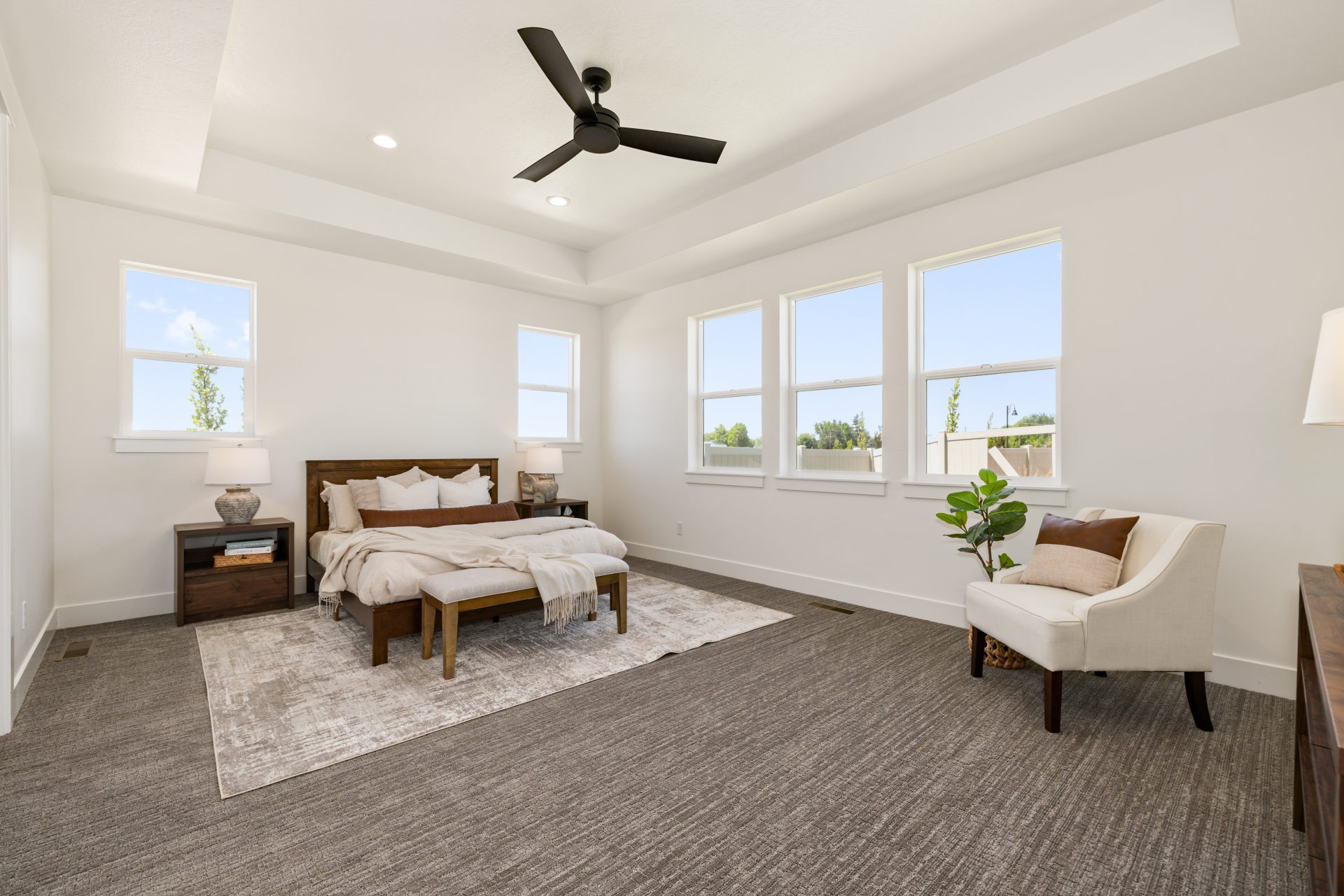 Carpeted bedroom with a tray ceiling, ceiling fan, and multiple windows bringing in abundant natural light. The room features a wood-framed bed with neutral bedding, a nightstand with a lamp, a bench at the foot, and an accent chair with a potted plant. T