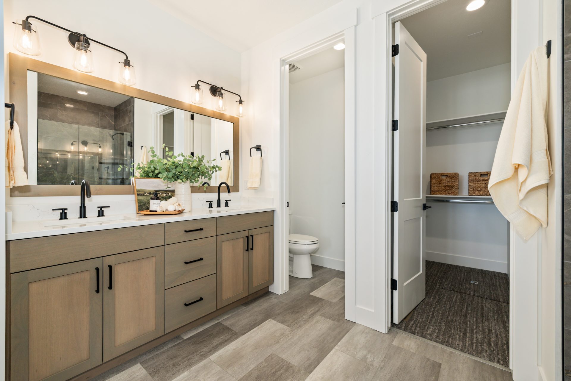 Bathroom with a dual-sink vanity, wood cabinets, and a light countertop, featuring a large framed mirror and sconce lighting above. The space includes a glass-enclosed shower, a separate toilet area, and a walk-in closet with built-in shelving, with tile