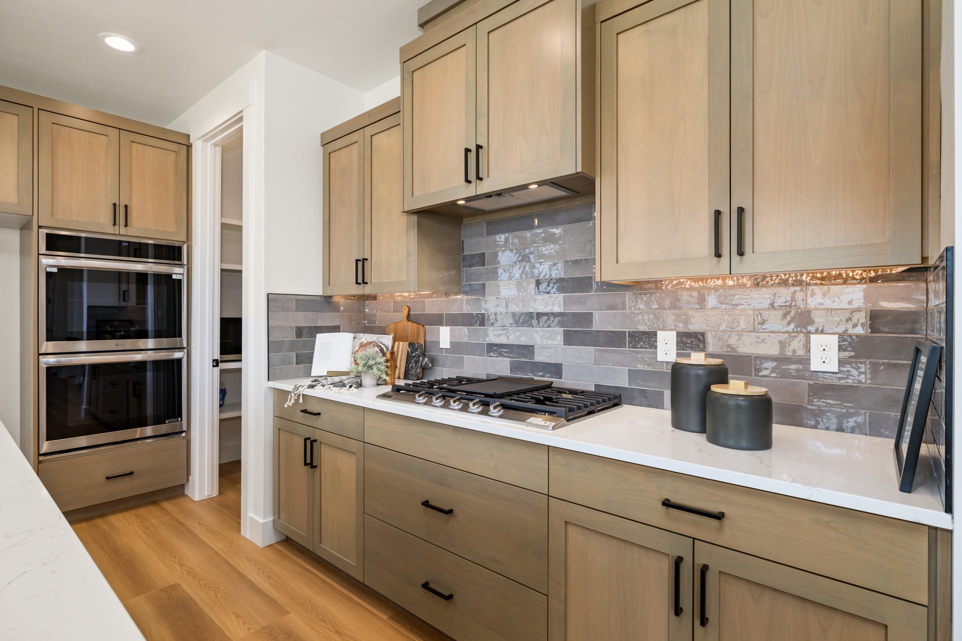 Kitchen with wood cabinets, marble-look countertops, and a gray tile backsplash. The space features a stainless steel gas cooktop, a built-in double oven, and under-cabinet lighting, with a walk-in pantry visible to the side. Light wood flooring and warm