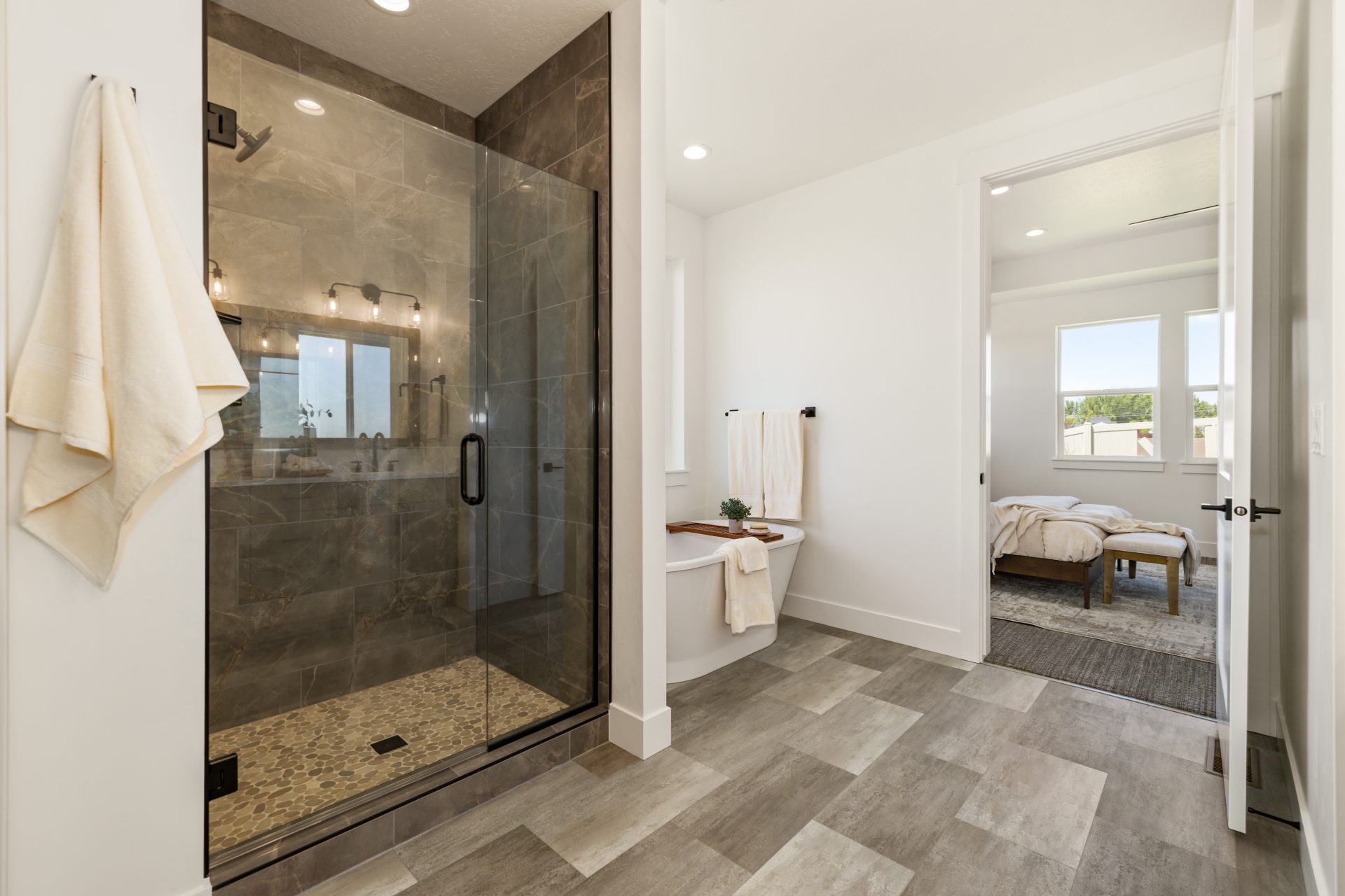 Bathroom with a glass-enclosed shower featuring dark tile surround and a built-in bench, beside a freestanding soaking tub. The space has tile flooring, light walls, and a towel hanging near the shower, with a view into the adjoining carpeted bedroom. Nat